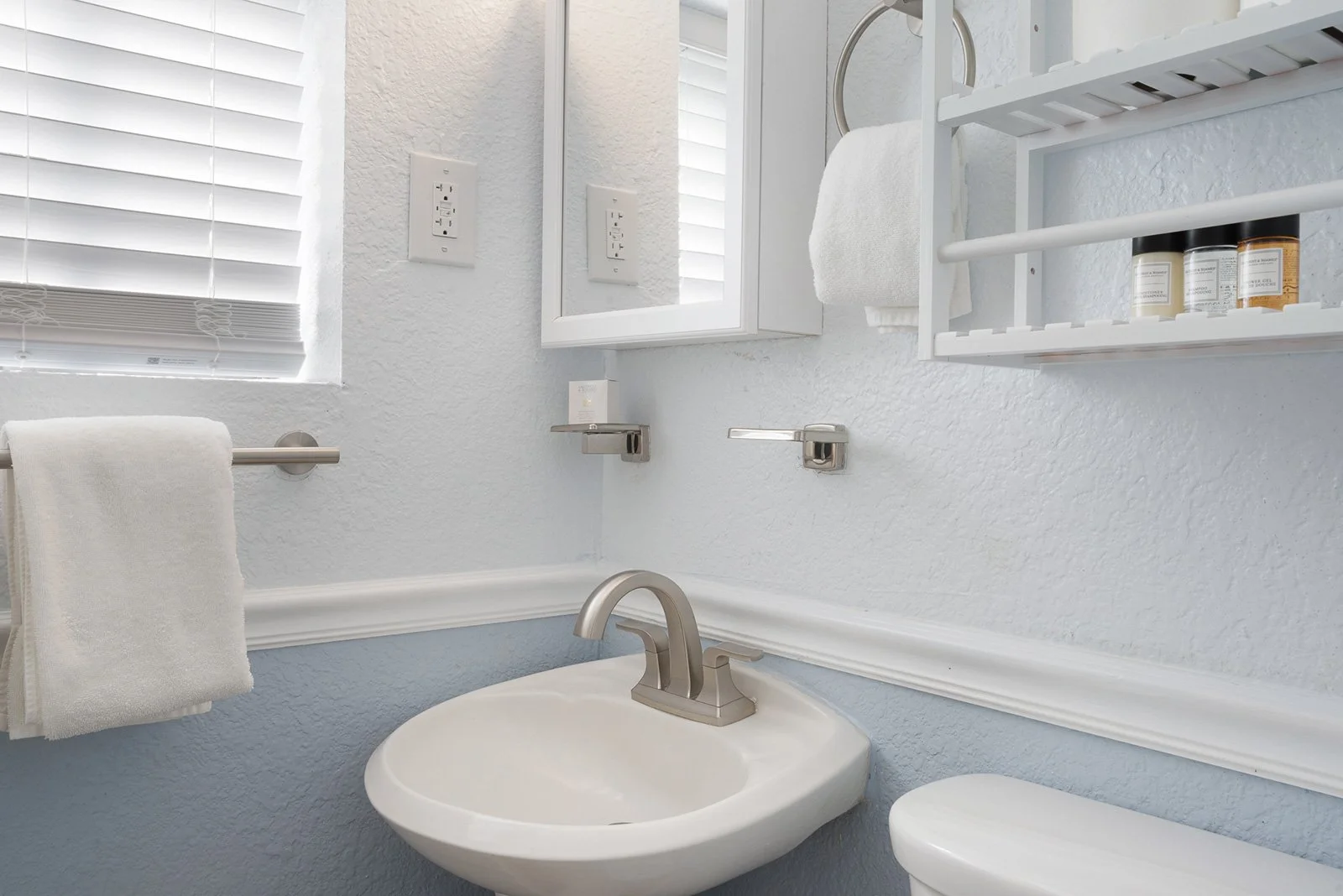 Bathroom with white textured walls, a small white sink with a silver faucet, a window with white blinds, a towel rail with a white towel, a wall-mounted mirror cabinet, and shelves with toiletries.