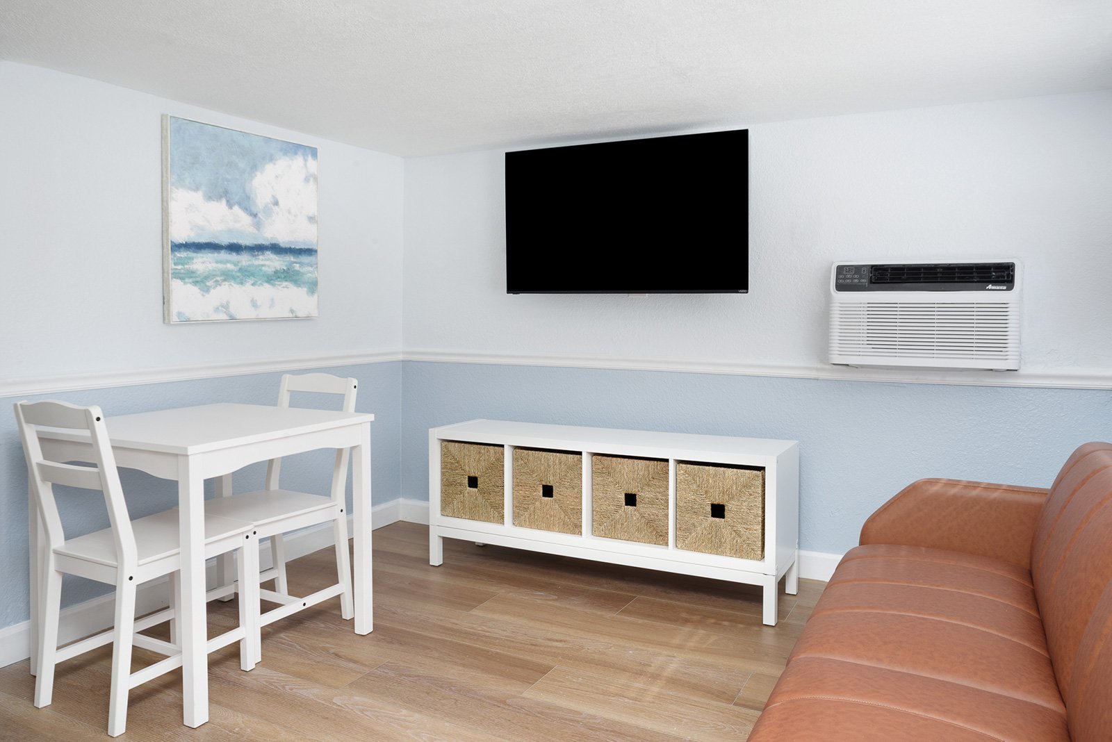 Living room with a white table and chairs, a white storage unit with wicker baskets, a tan couch, a wall-mounted TV, an air conditioning unit, and a painting of the ocean on white and light blue walls.