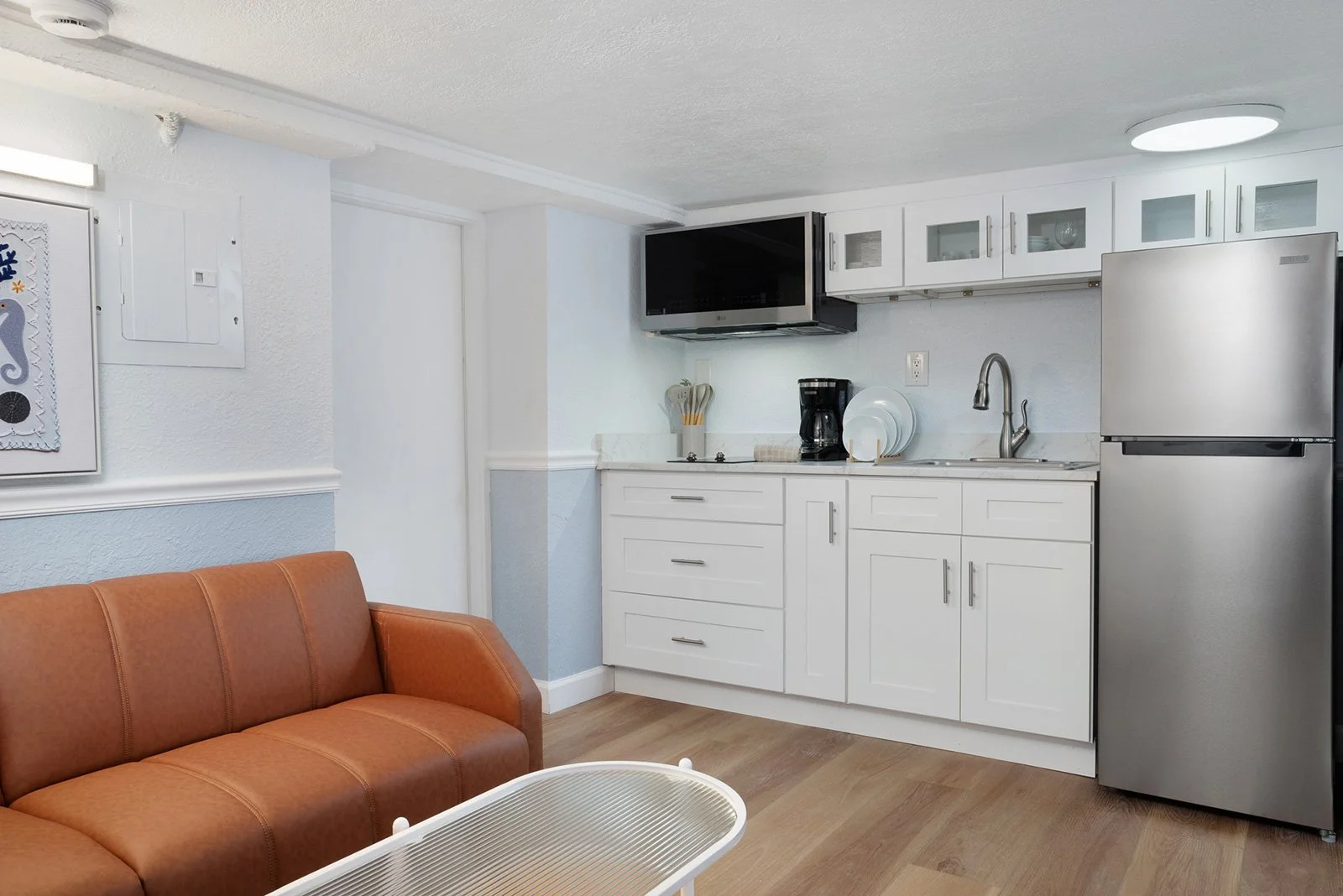 Living area with a brown sofa, white coffee table, white kitchen cabinetry, stove, coffee maker, dishes, and a refrigerator in a small apartment or hotel room.