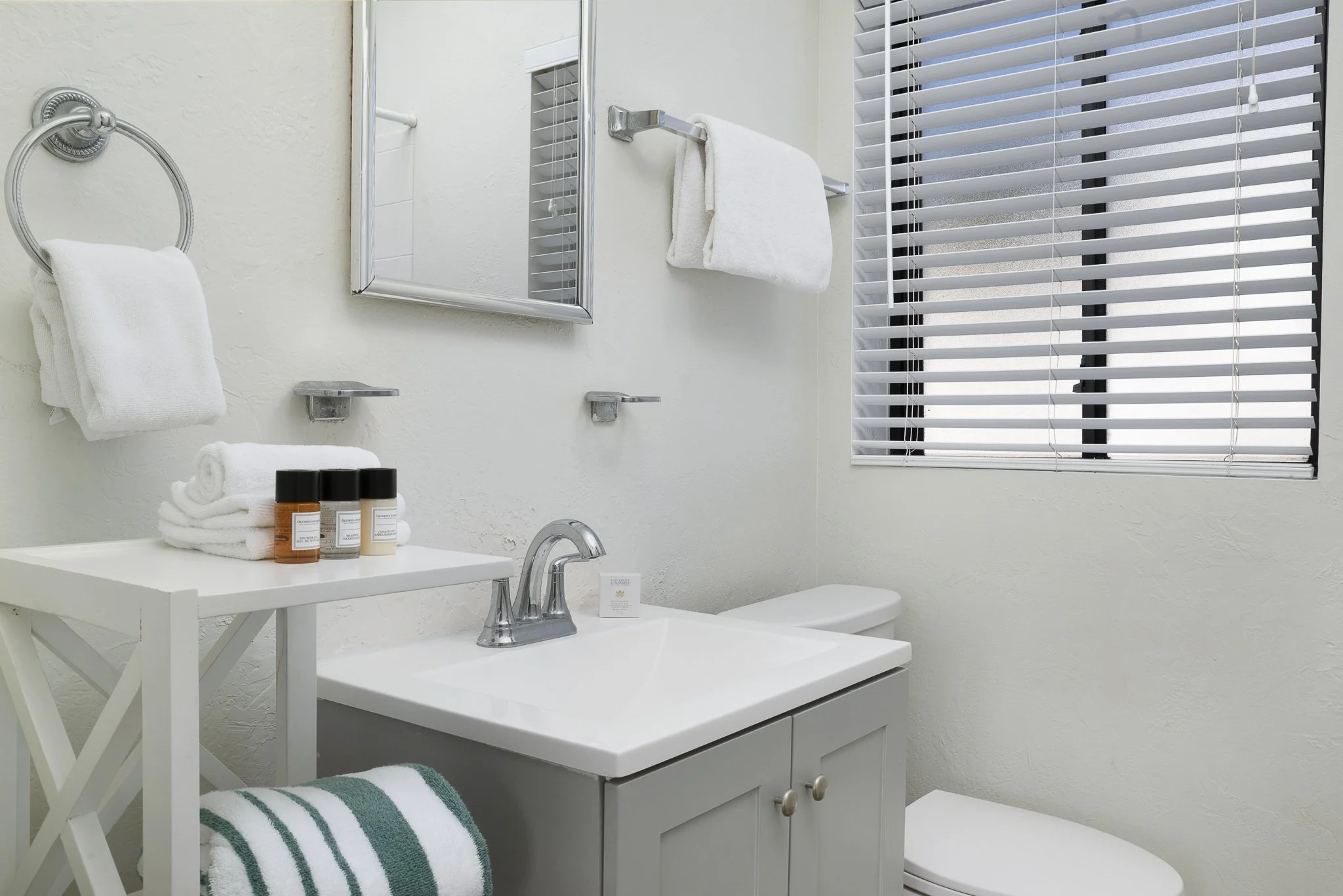 Clean white bathroom with a sink, mirror, toilet, and window with blinds. Towels and toiletries are neatly arranged.