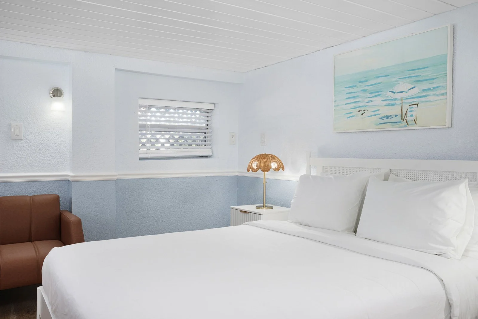 A beach boutique hotel bedroom with white walls and ceiling, a bed with white bedding, a nightstand with a lamp, a small brown sofa, a window with blinds, and a beach-themed painting on the wall at Sabal Palms inn.