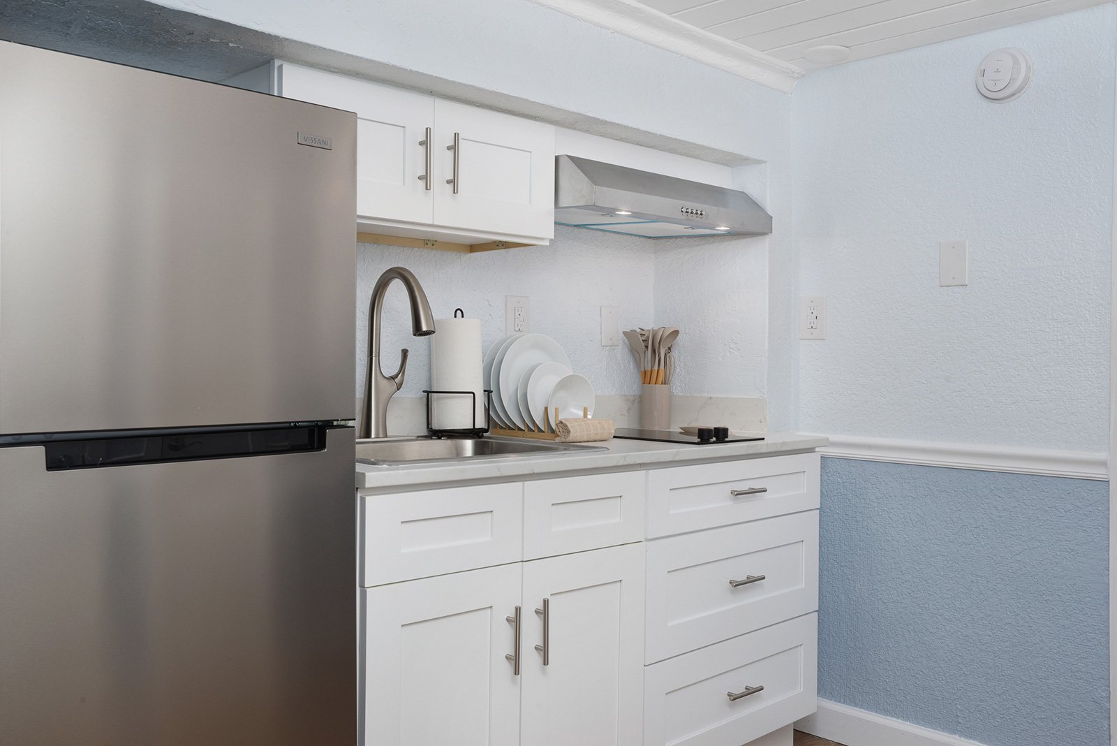 Modern kitchen with stainless steel refrigerator, white cabinets, a sink with a faucet, a paper towel holder, a set of white plates, a utensil holder, and a stovetop with two burners, with a vent hood and wall outlets.