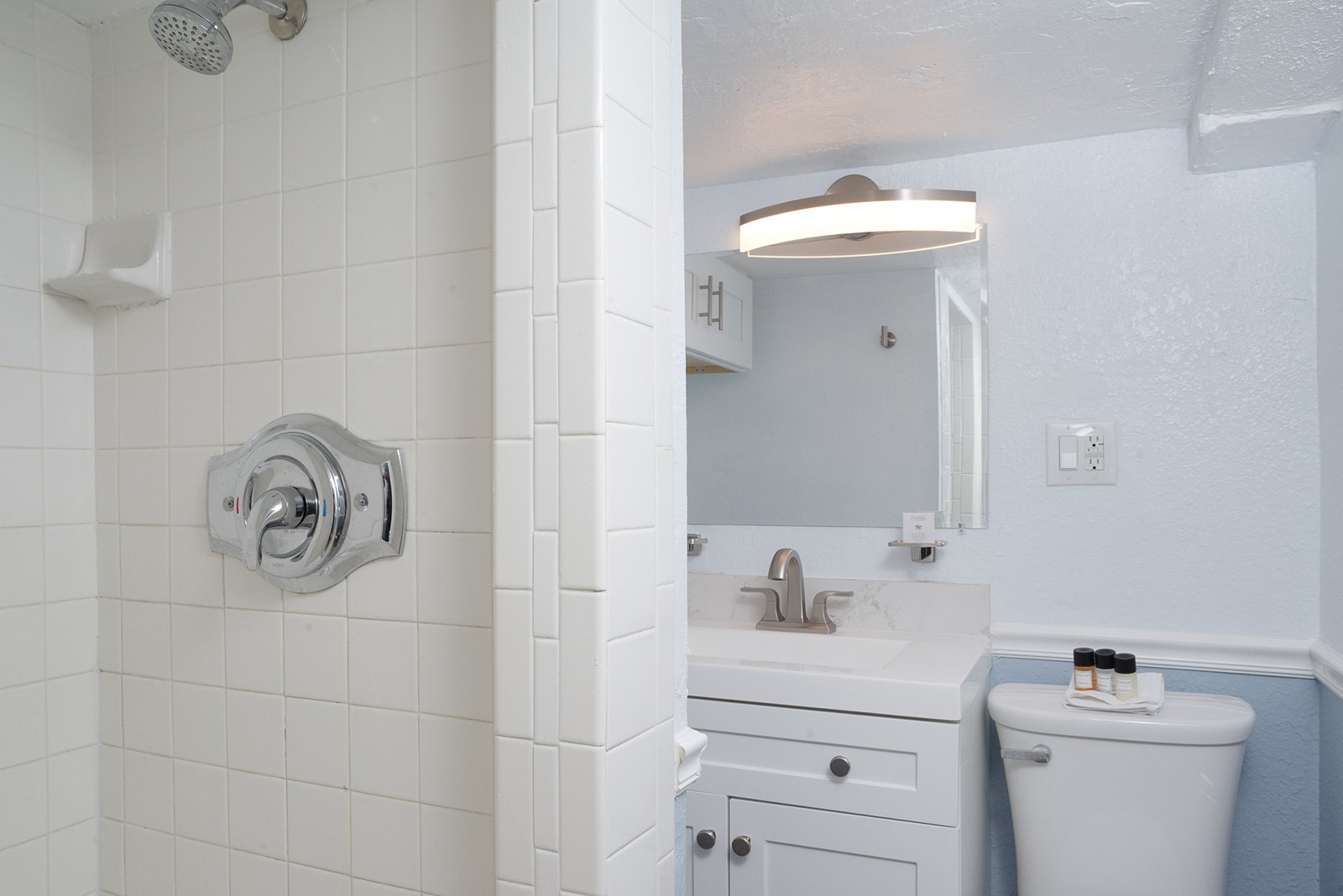 Bathroom with a shower, white tiles, and a vanity with a mirror. The toilet has small bottles of toiletries on top.