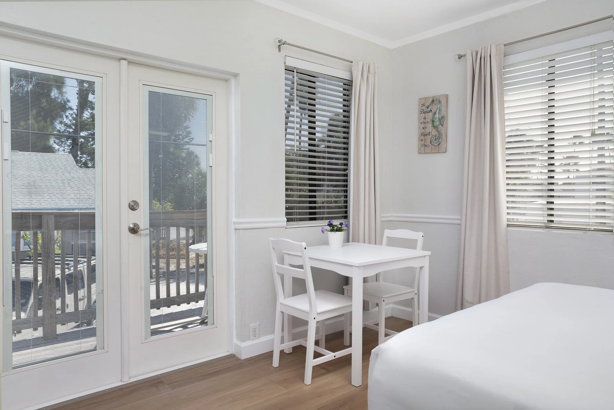 A bright hotel room interior featuring white walls, two large windows with opened blinds and light curtains, a glass door with blinds leading to a balcony, a white table with two matching chairs and a potted plant on top, partially visible bed with w