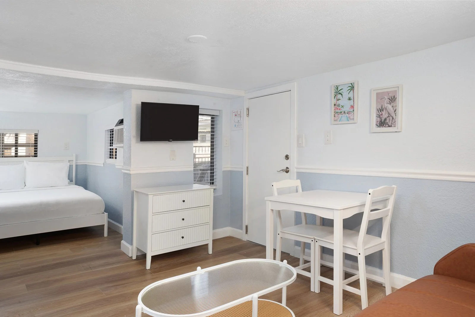 Interior of a light blue and white hotel room with a bed, TV, small dining table and chairs, a couch, and wall art featuring tropical themes.