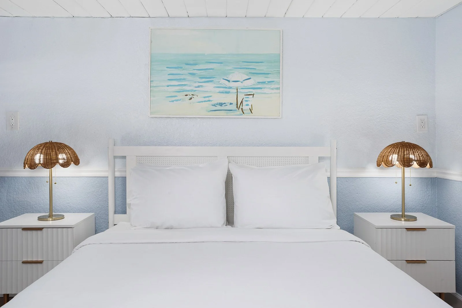 A neatly made bed in a beach boutique hotel with white pillows and bedding in a room with white-paneled walls. On each side of the bed are white nightstands with wicker lamps. A painting of a beach scene hangs above the bed at Sabal Palms inn..