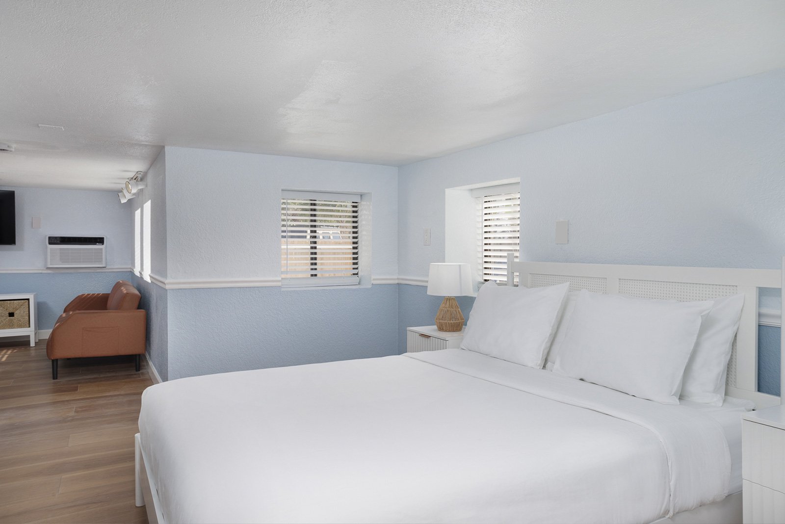 A cozy bedroom with a white bed, white pillows, a nightstand with a lamp, light blue walls, and windows with white blinds, with a seating area and air conditioning unit in the background.