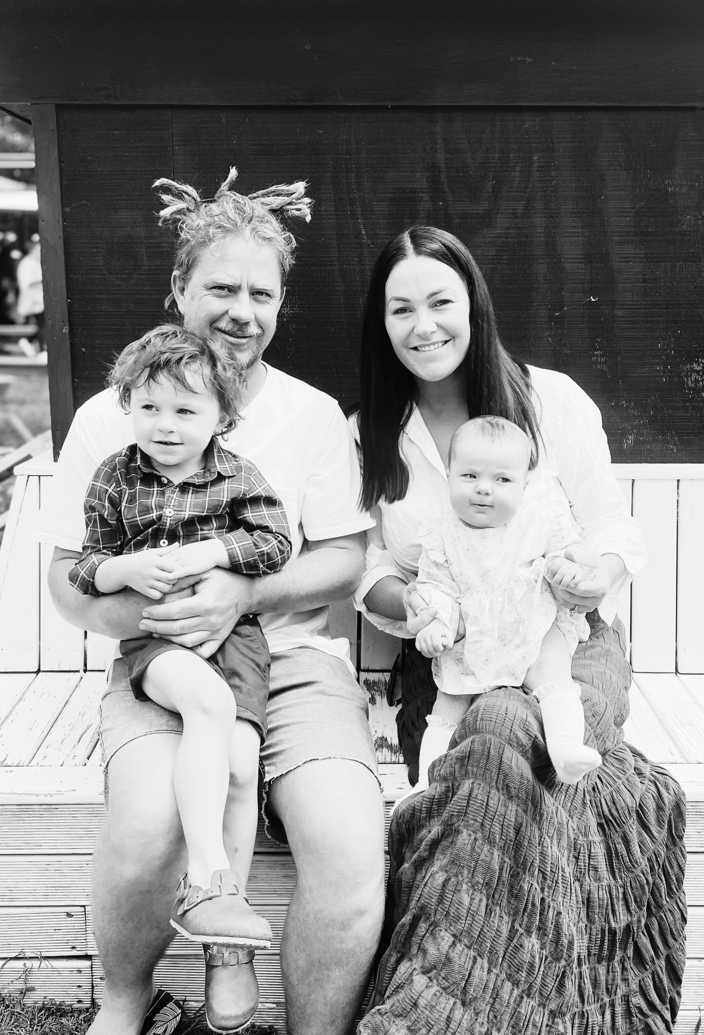 Black and white image of a family sitting on a bench; two adults holding a young child and a baby.