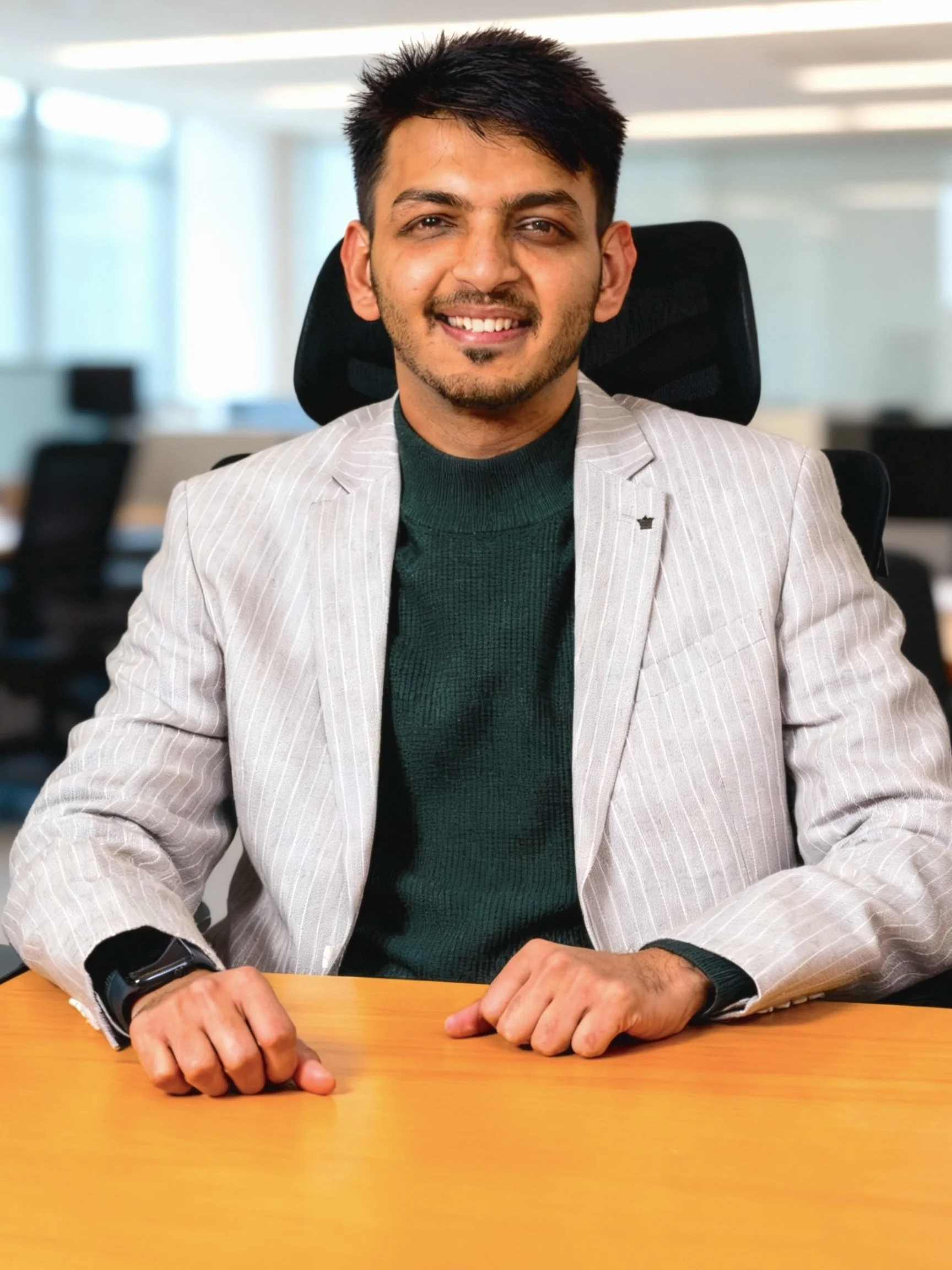 A smiling man with short dark hair, wearing a light striped blazer and dark green turtleneck, seated at a wooden table in a modern office.