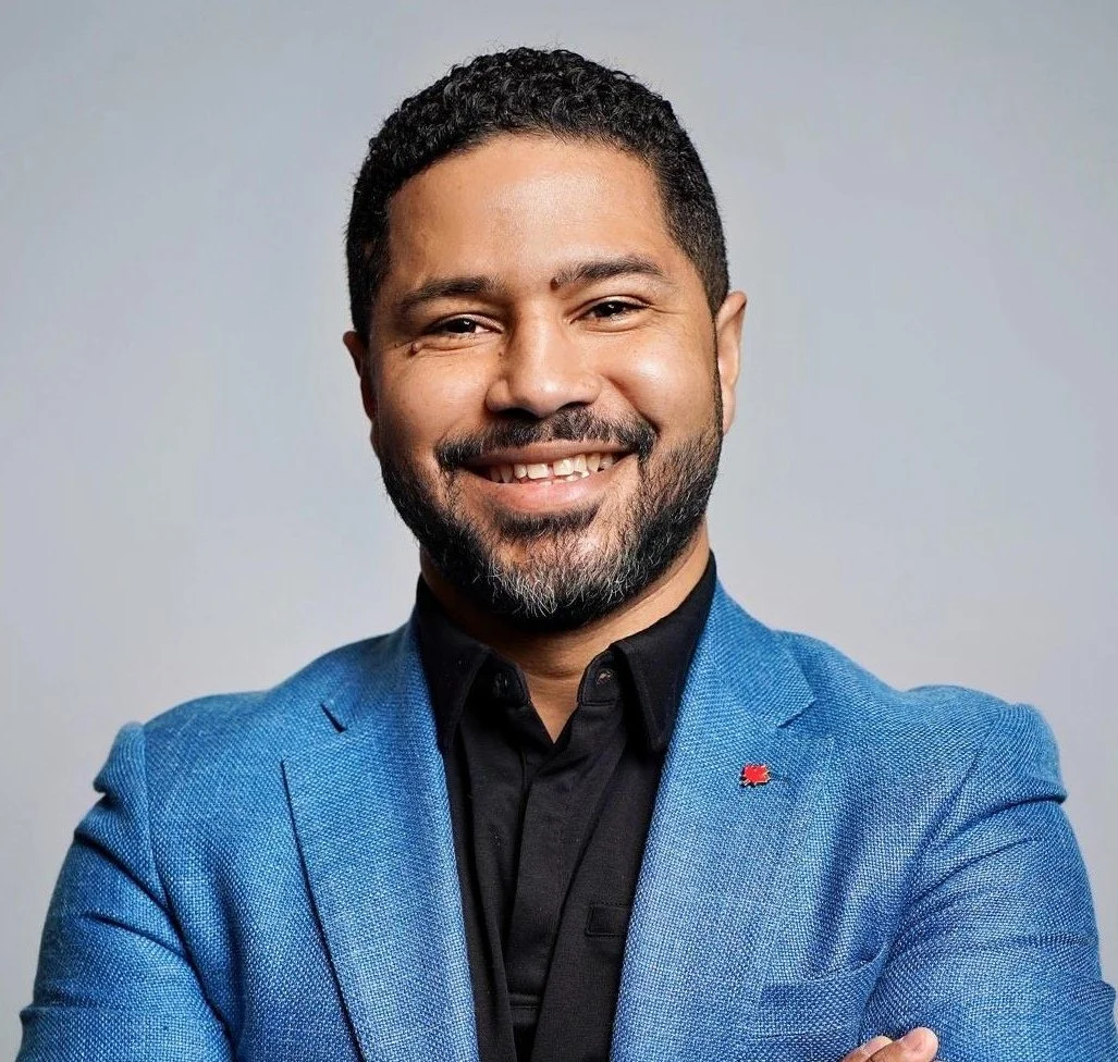 A smiling man in a blue blazer and black shirt, arms crossed, against a gray background.