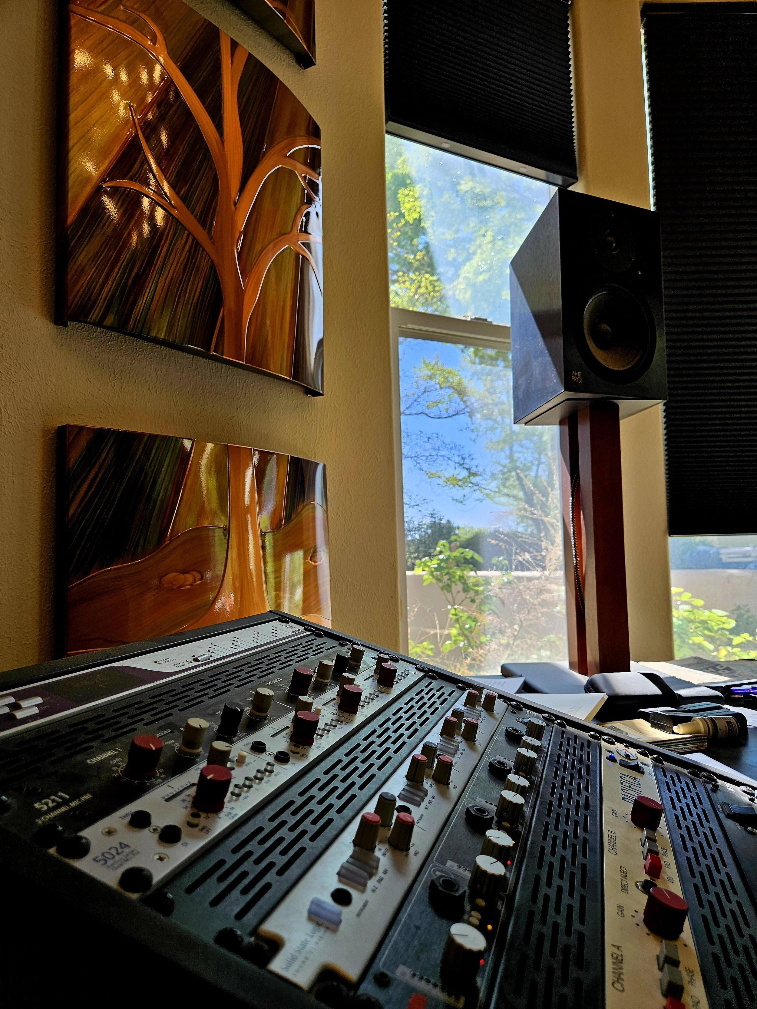 Recording studio with audio mixer, studio monitor speaker, and soundproof window showing trees outside.