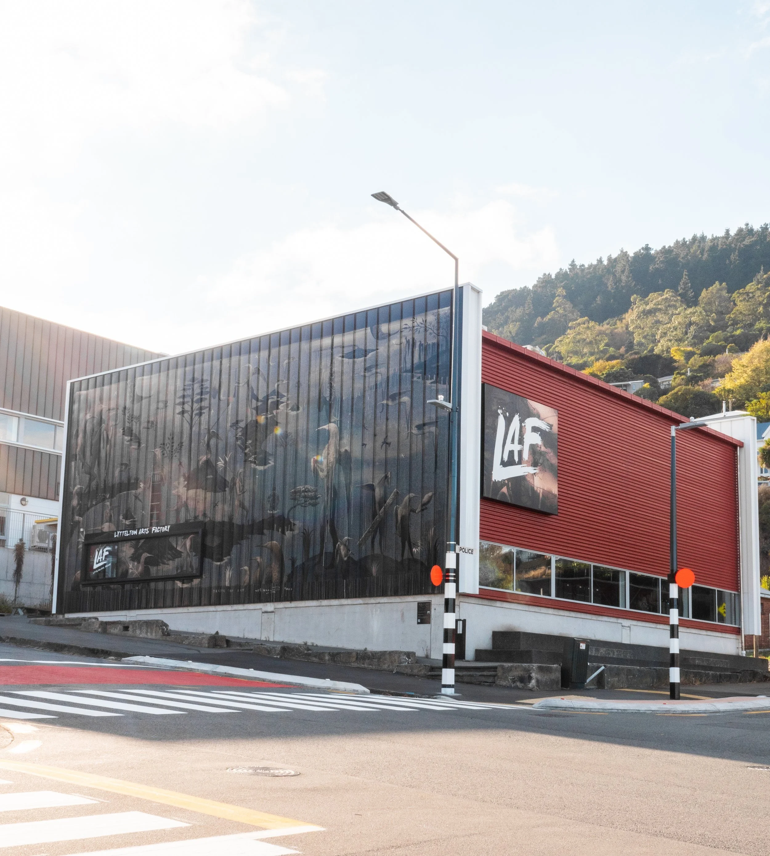 A modern building with a large mural of birds on the black metal exterior and a red facade. There is a sign that reads "Lyttelton Arts Factory" and a smaller sign "IAF." A street with a crosswalk and poles with orange reflectors is in the foreground. Hills with trees are in the background.