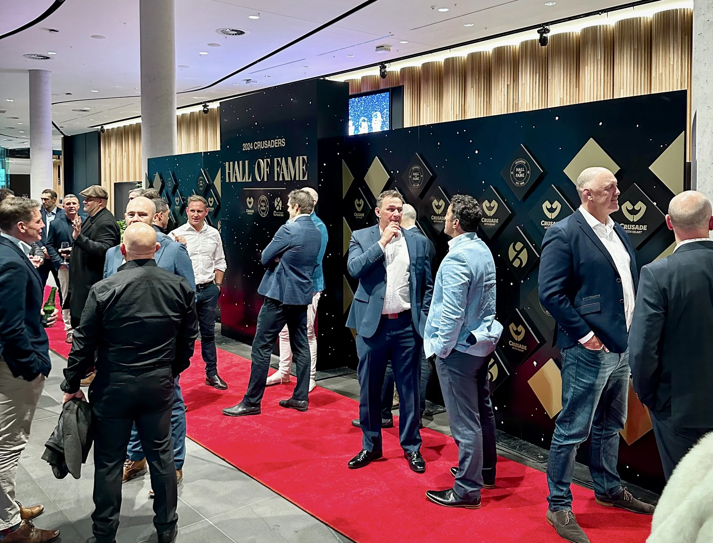 A group of men in suits and business casual clothing are gathered at an indoor event with a red carpet, socializing in front of a backdrop that reads '2024 Crusaders Hall of Fame.' The backdrop features black and gold design elements and logos.