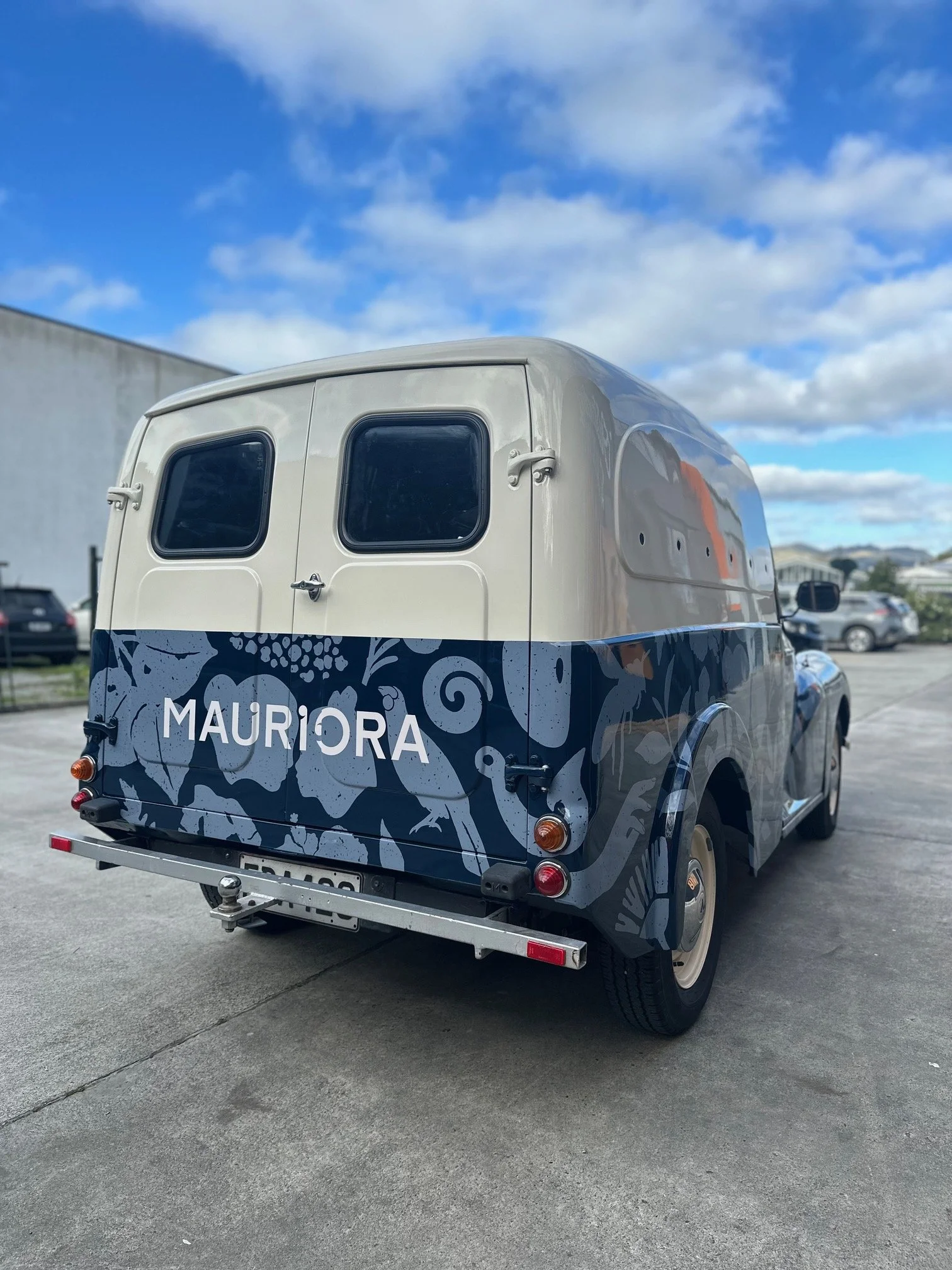 Vintage delivery van with a dark blue and white floral pattern and the word "MAURIORA" on the back, parked outdoors under a partly cloudy sky.