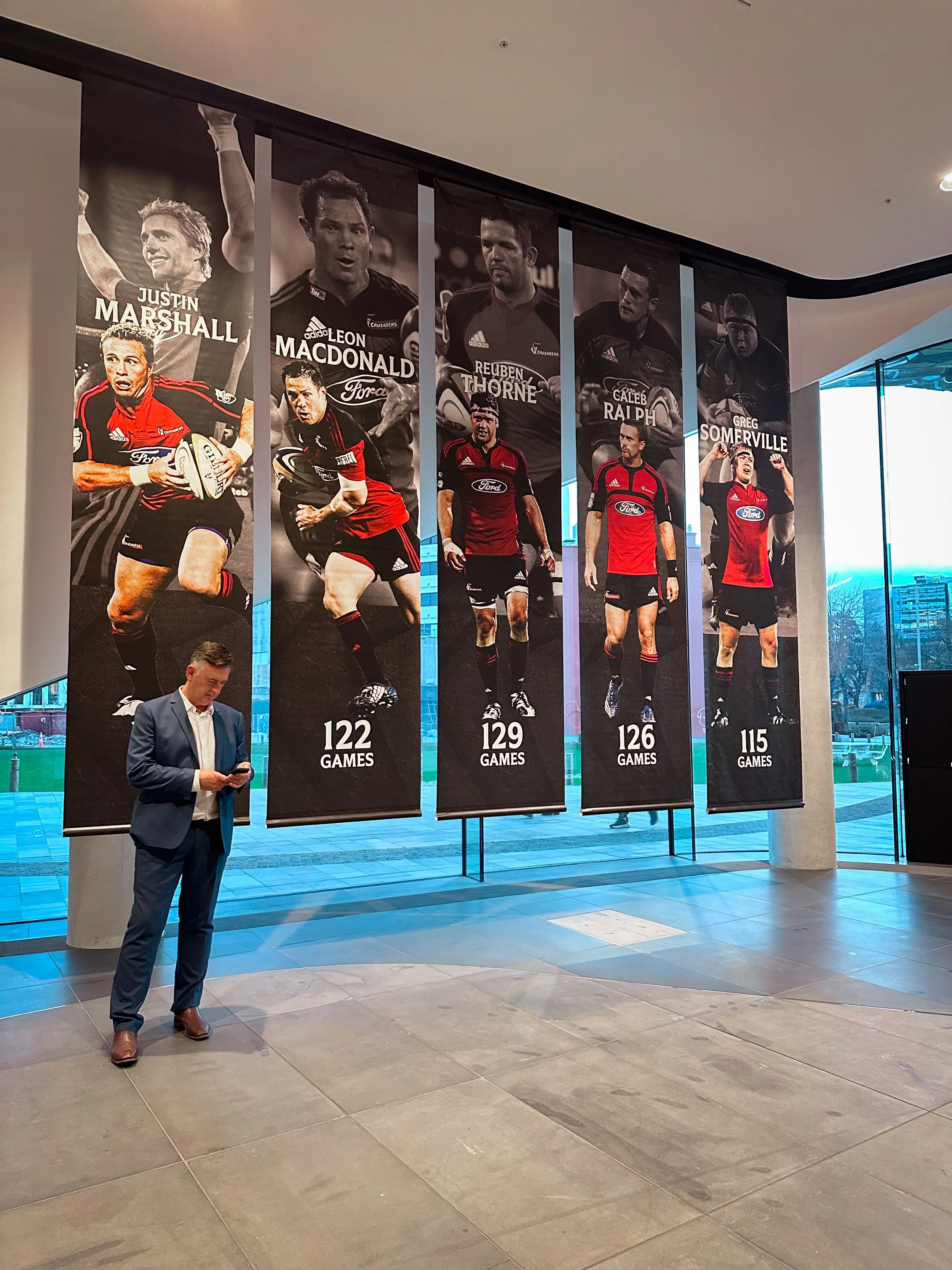 Interior of a building with a man in a suit looking at his phone, and large vertical banners hanging from the ceiling showcasing rugby players and their number of games played, labeled from left to right as 122, 129, 126, and 115 games.