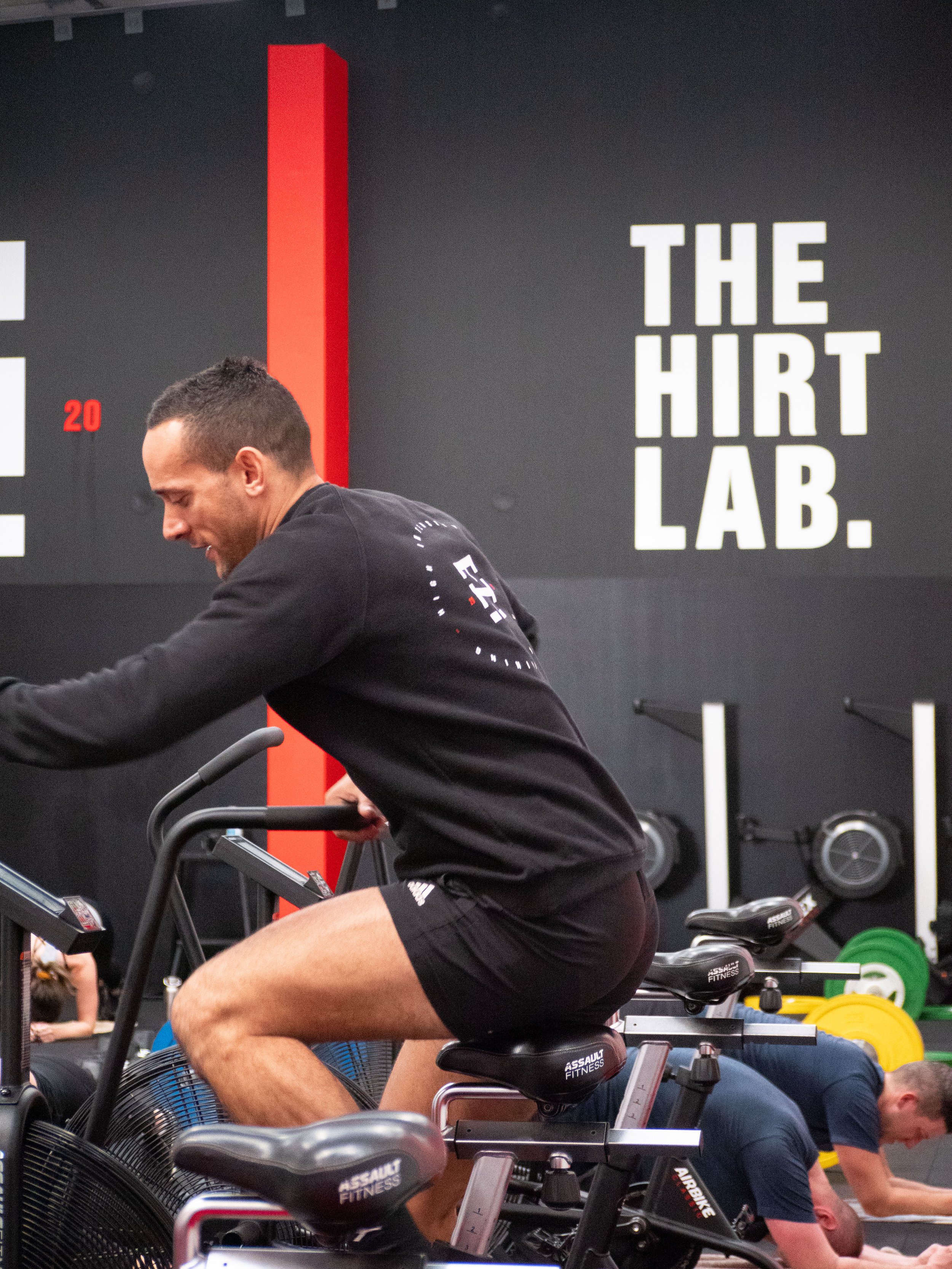 A man riding a stationary exercise bike in a gym with a black wall that has a large red vertical bar and the text 'THE HIRT LAB.'