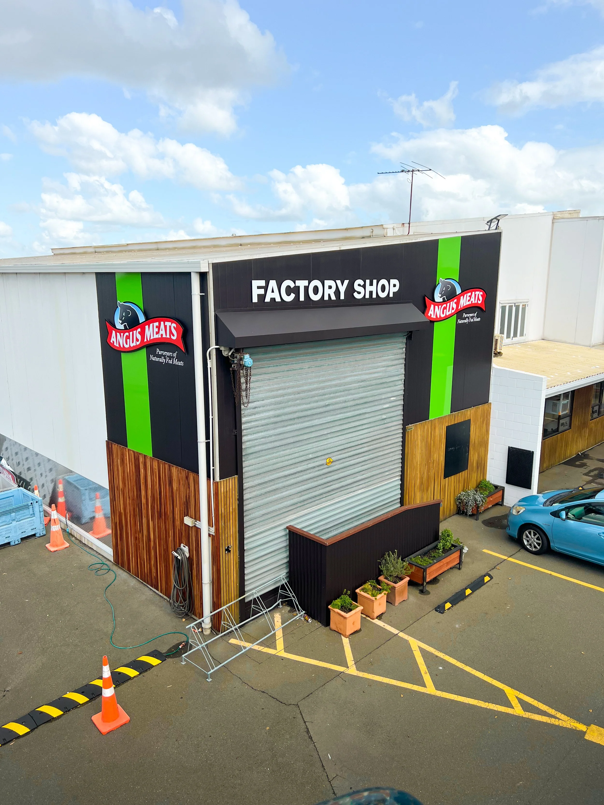 Exterior of a factory shop building with a closed metal roll-up door, black and green signage, and a parking lot with orange cones and potted plants.