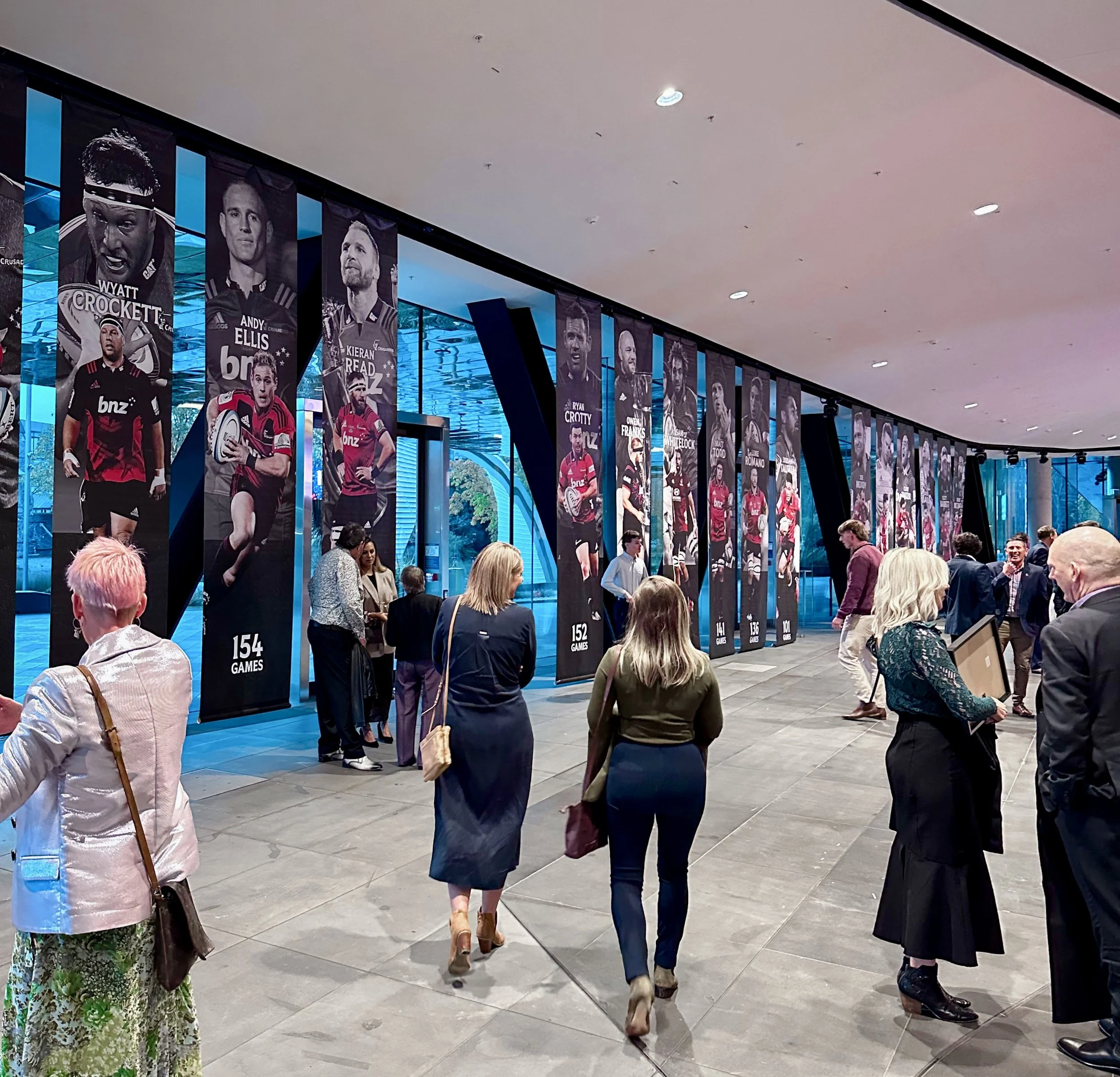 People walking and conversing inside a modern building with large banners displaying rugby players and statistics. The banners feature photographs of rugby players with their names, team logos, and game counts.