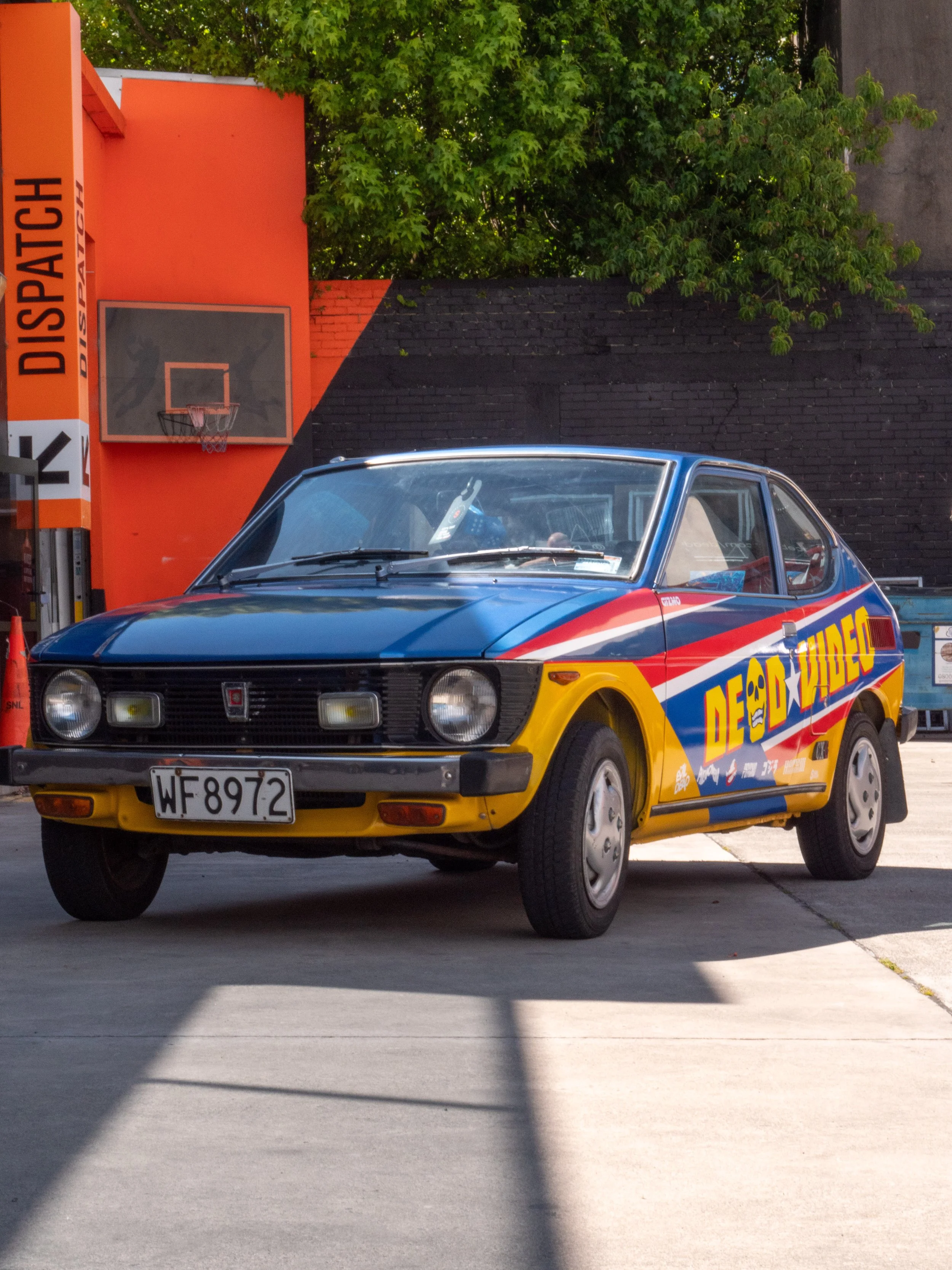 A vintage sports car with colorful graphics and the words 'DEAD RIDER' painted on its side, parked on a concrete surface near a mini basketball hoop and a black brick wall, with green trees in the background.