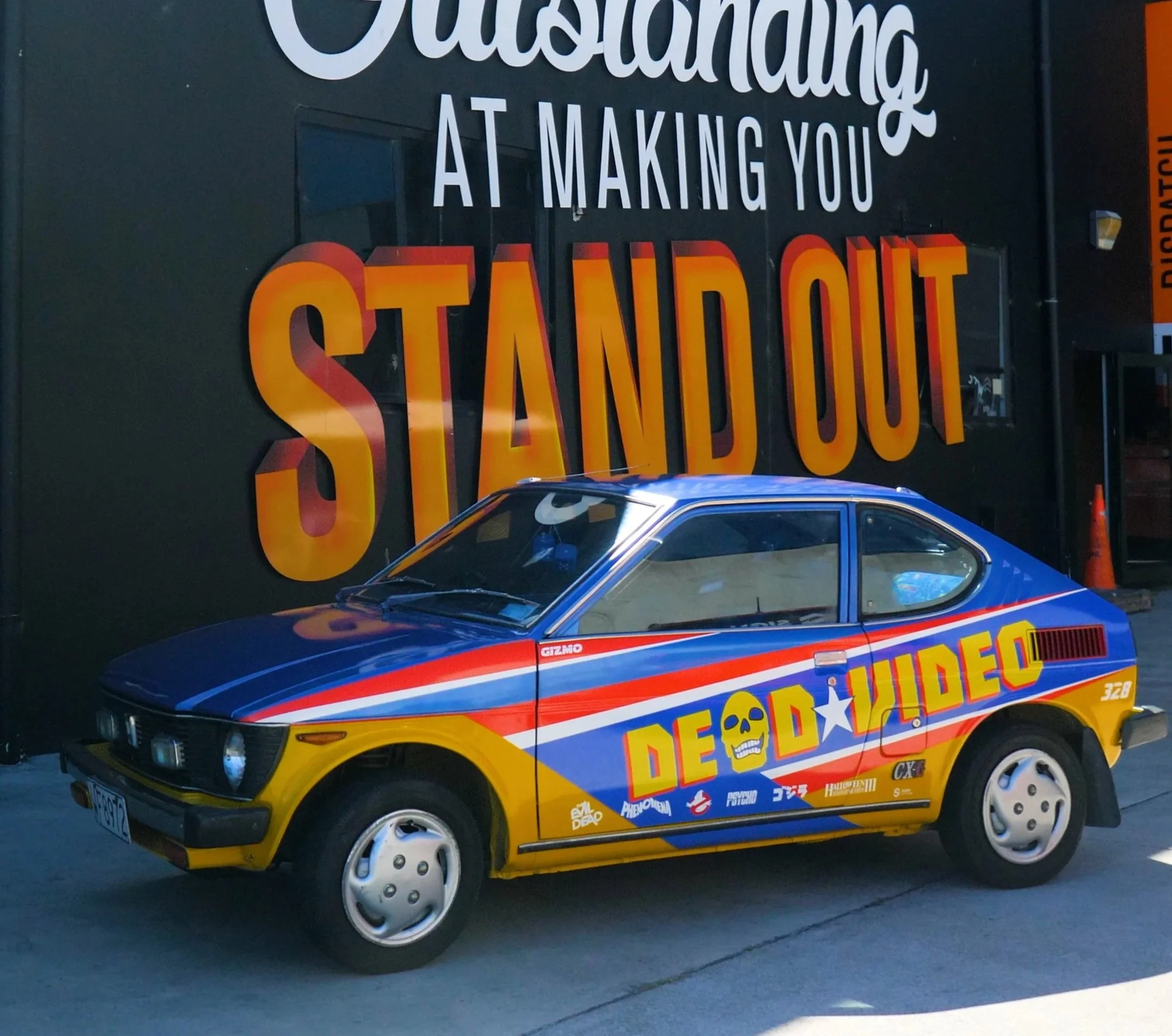 A vintage IROC-Z Camaro car painted with colorful decals and graphics, parked in front of a building with a sign that reads, 'Outstanding at Making You STAND OUT.'
