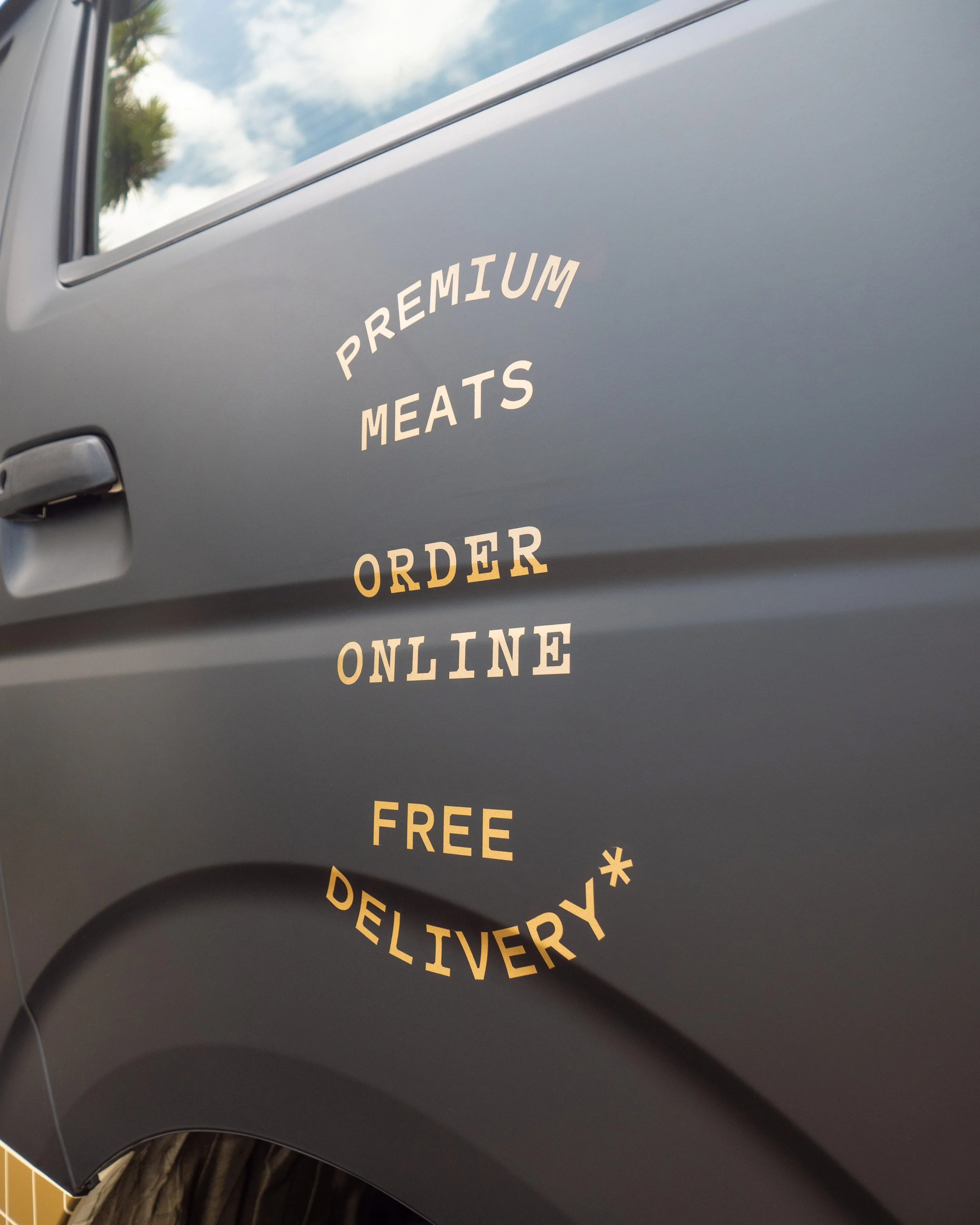 Text on the side of a gray vehicle advertising premium meats for online ordering with free delivery, with a small asterisk.