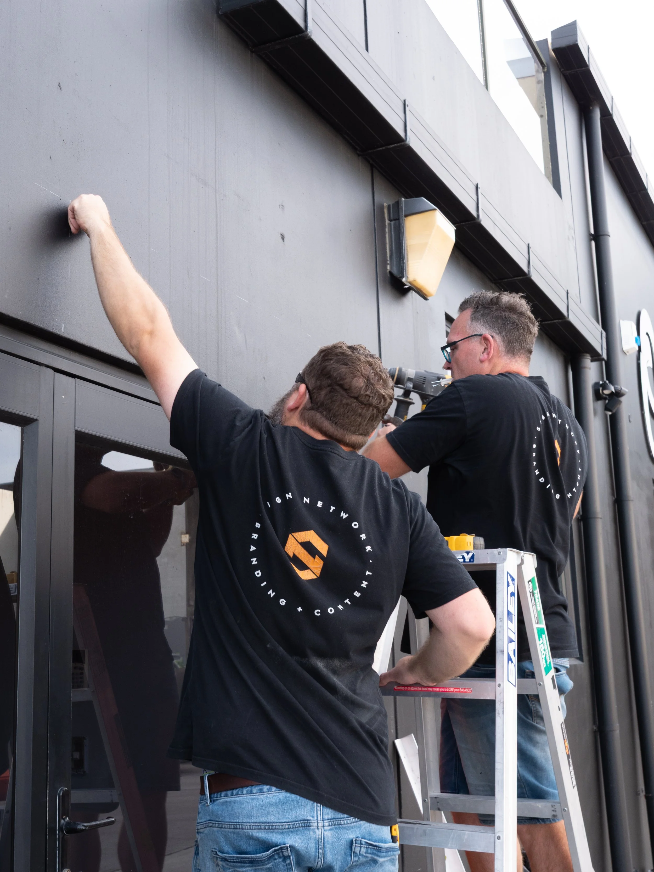 Two men working on the exterior of a building, one on a ladder and the other standing, both wearing black T-shirts with a logo and text. One man is drilling into the wall while the other observes.