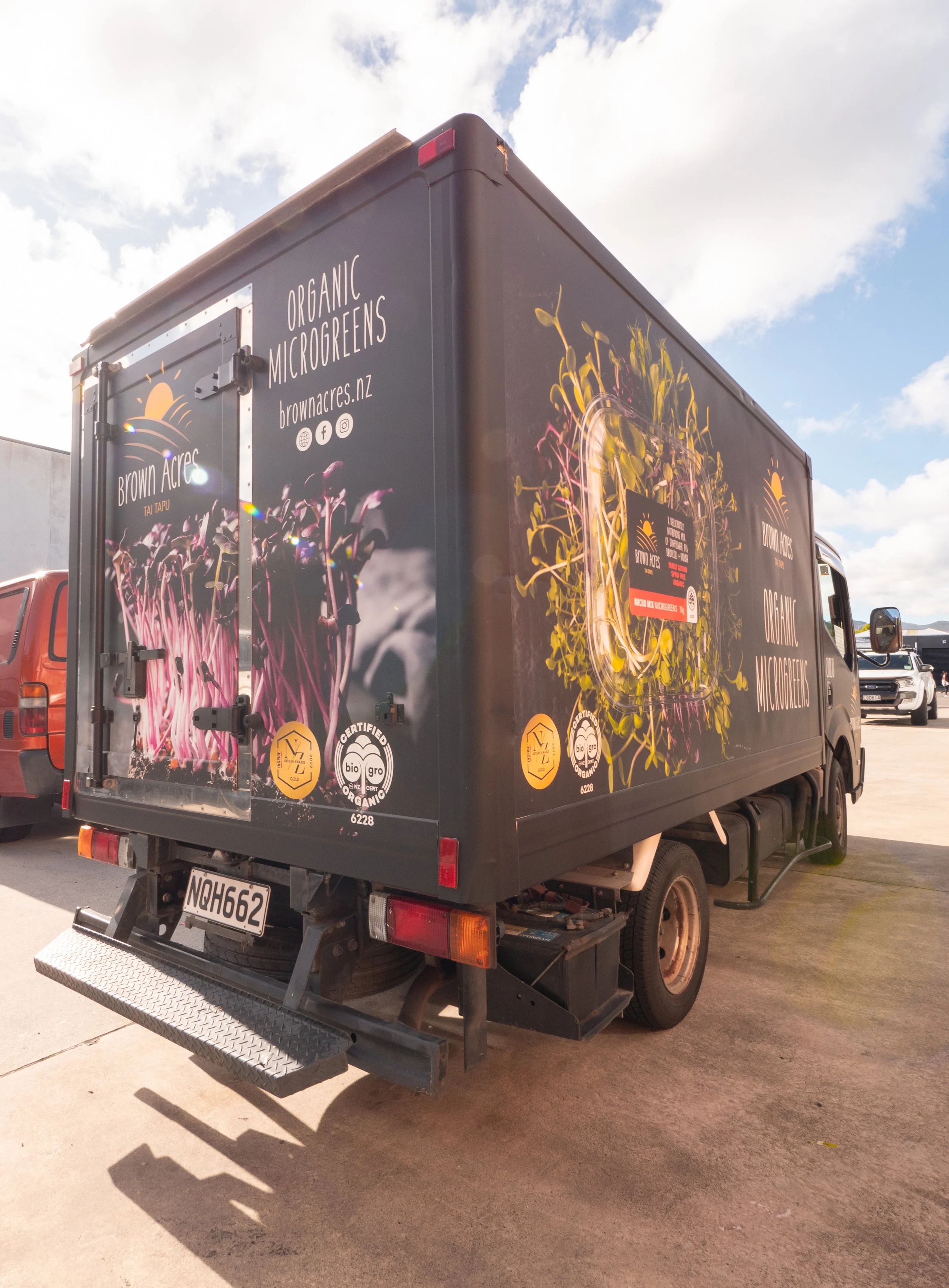 A black delivery truck with colorful graphics and text promoting organic microgreens from Brown Acres, parked on a concrete lot under a partly cloudy sky.
