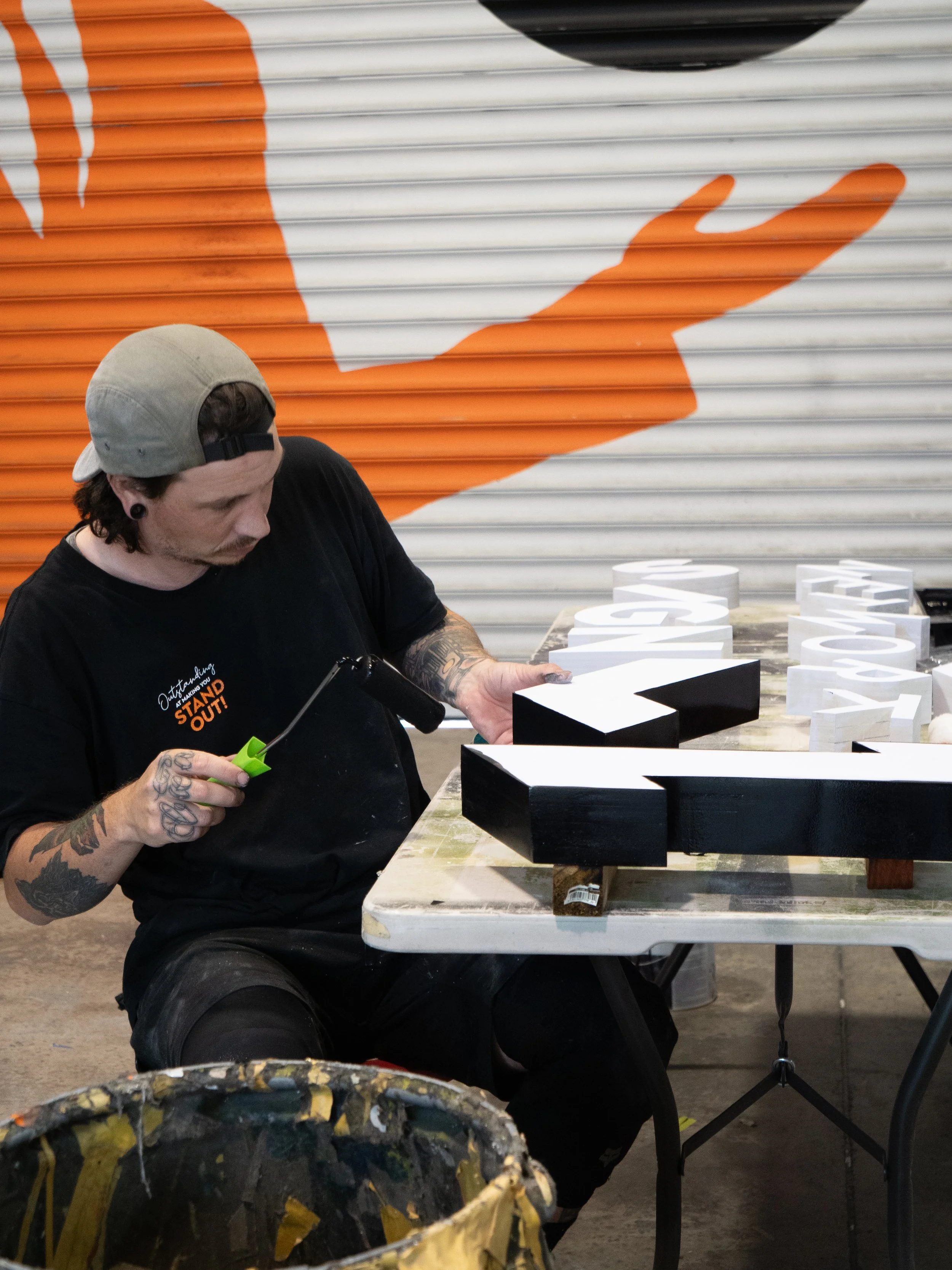 A man wearing a black T-shirt and a gray backward baseball cap is painting black and white wooden letters on a table. There is a large orange and white hand graphic painted on a metal shutter in the background.
