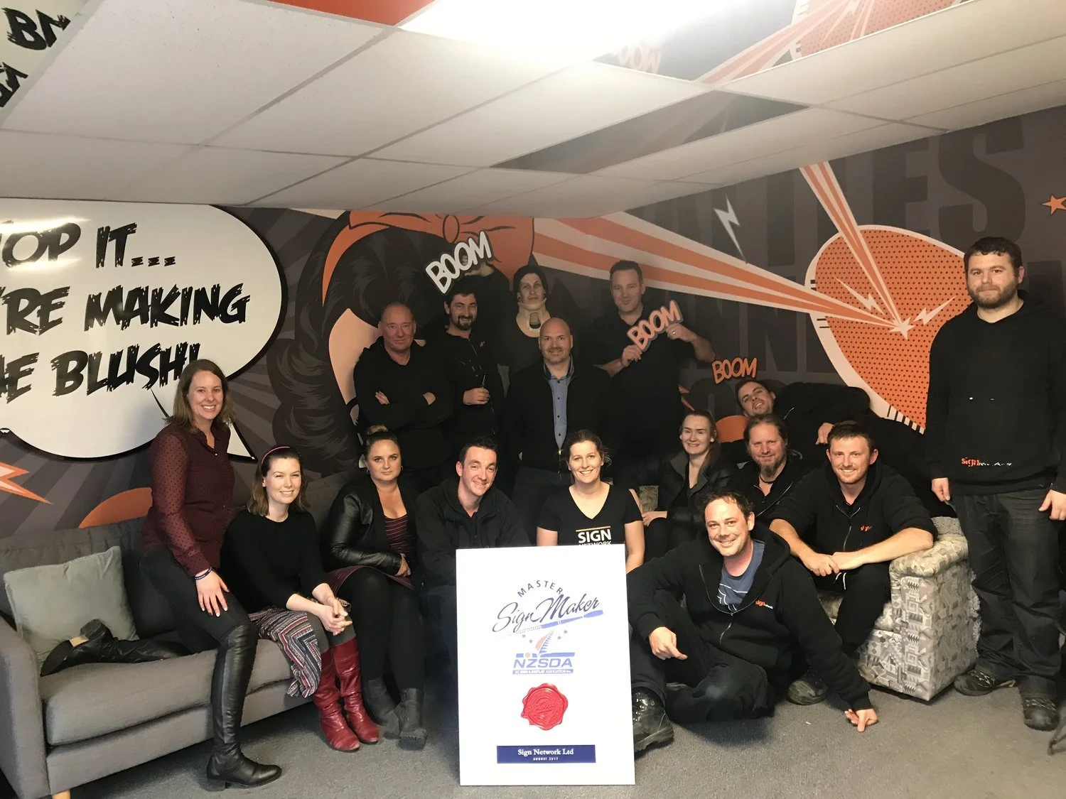 Group of people posing together in front of a comic-style wall mural with speech bubbles and explosion graphics, holding a sign that says 'Sign Maker Master' with a red wax seal, at an indoor event.