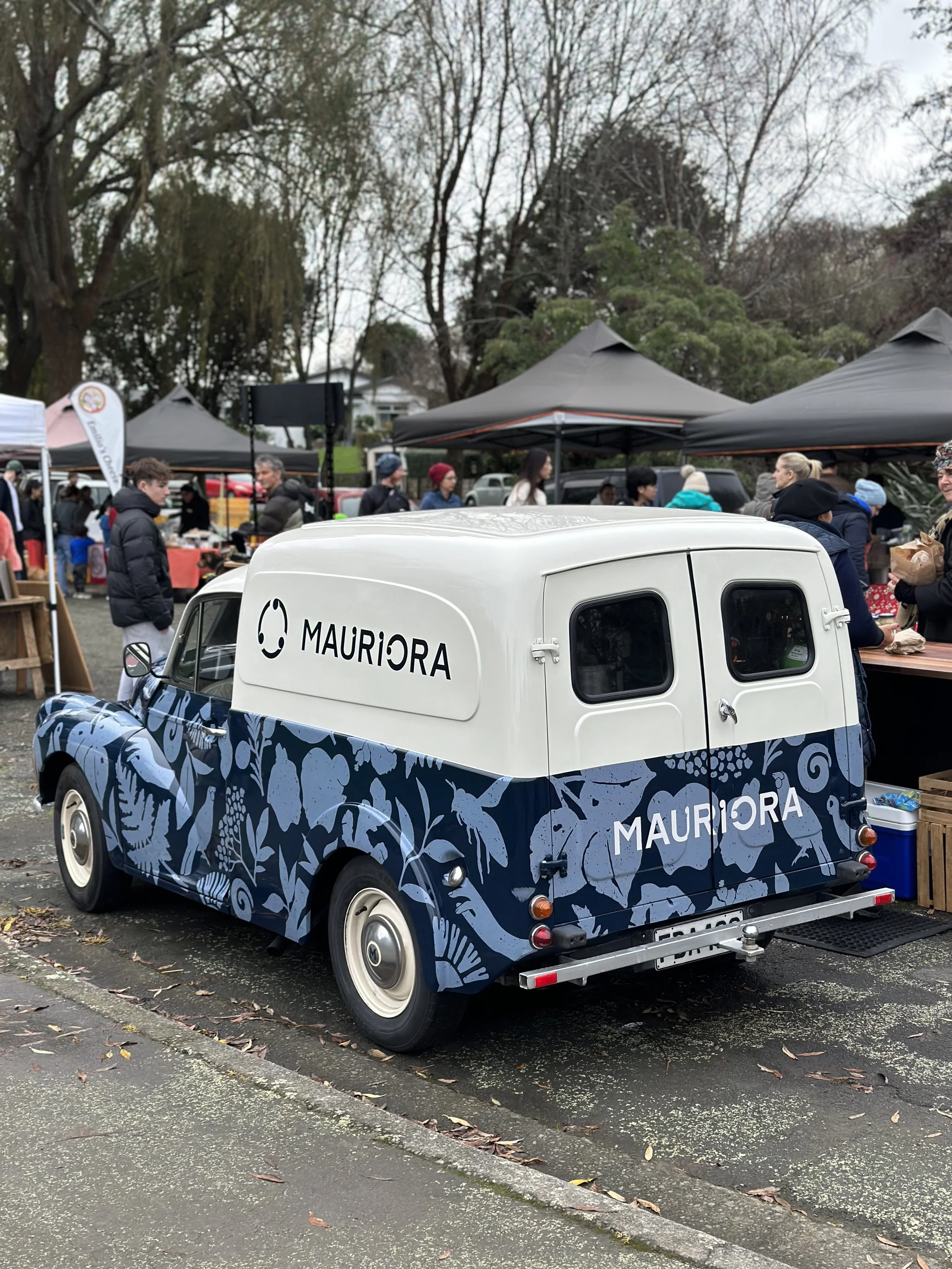 A vintage van with a white top and navy bottom decorated with floral and animal patterns, parked at an outdoor market, with tents and people in the background.