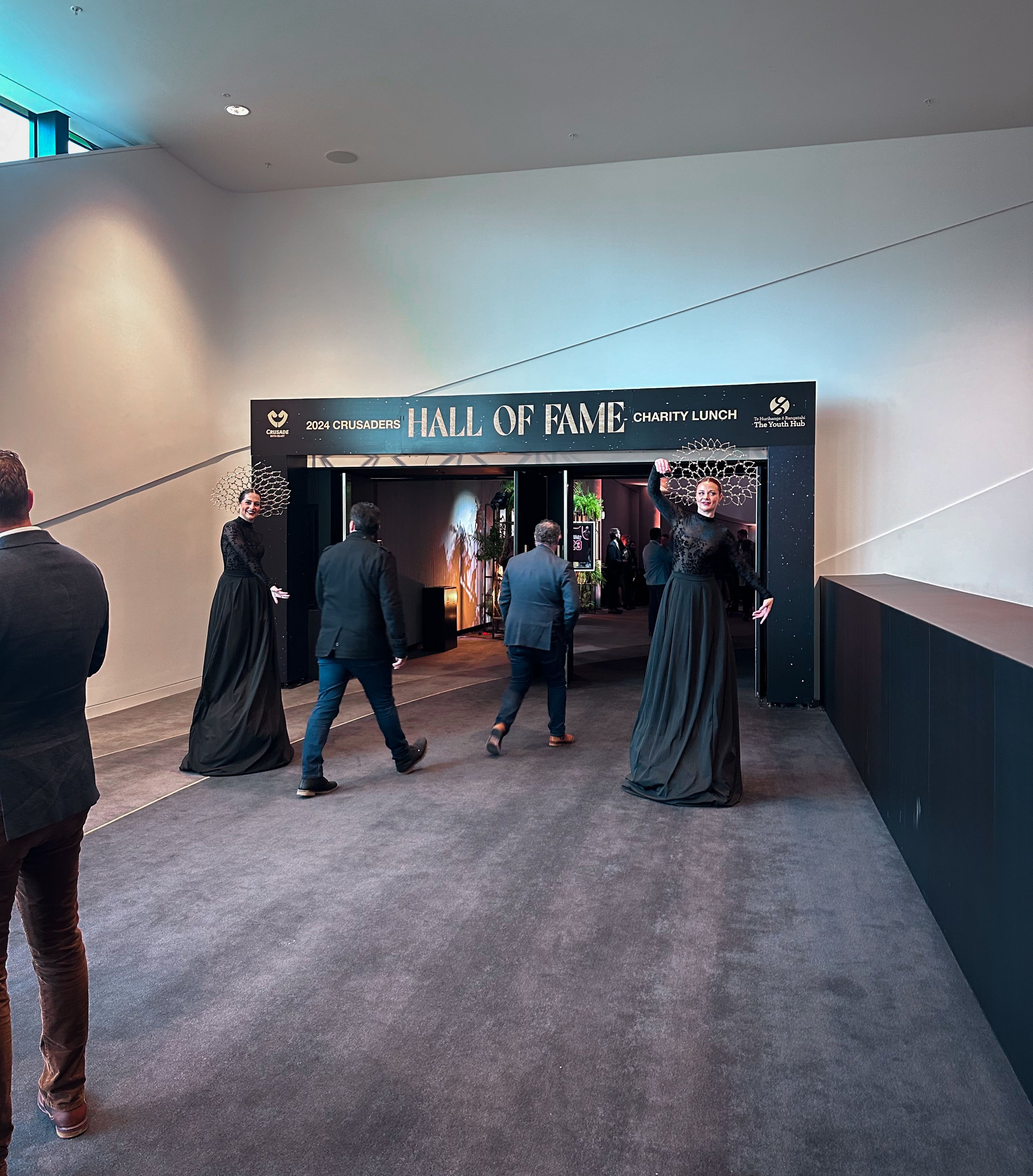 Entrance to the 2024 Crusaders Hall of Fame charity lunch event with two tall fashionably dressed women standing on tall stilts on either side of the entrance, and several people walking toward the entrance in a modern, well-lit indoor venue.
