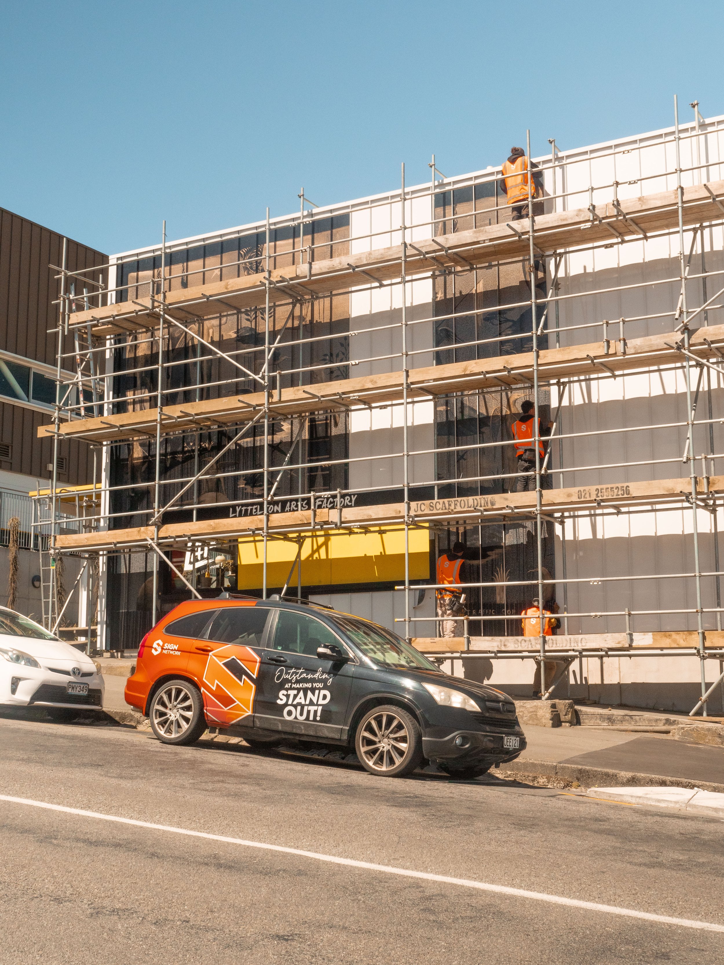 Construction workers on scaffolding work on the exterior wall of a building, with a branded vehicle parked in front and a clear blue sky overhead.
