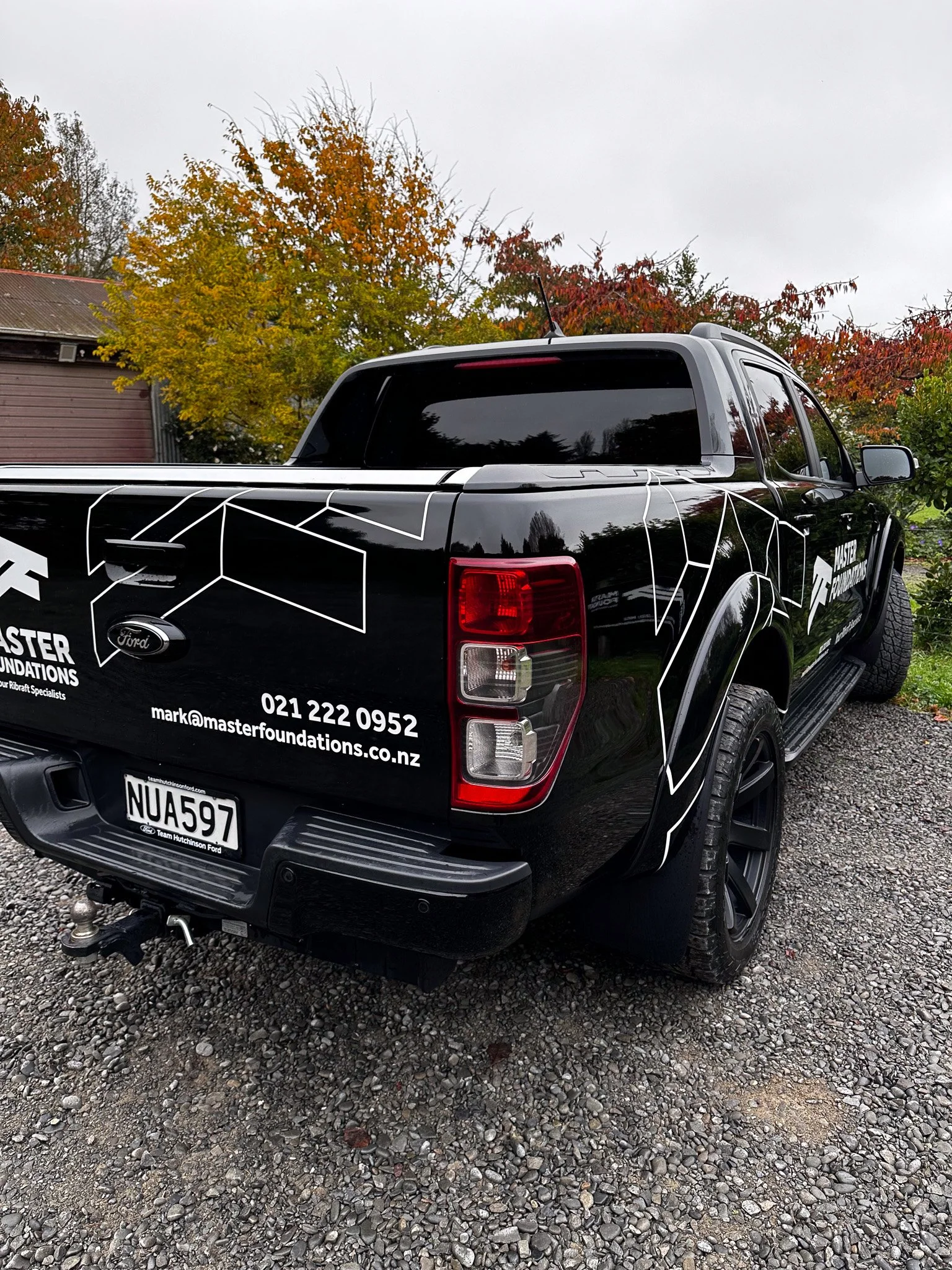 Black pickup truck with white geometric line art and the logo 'Master Foundations' parked on gravel, with autumn trees in the background.