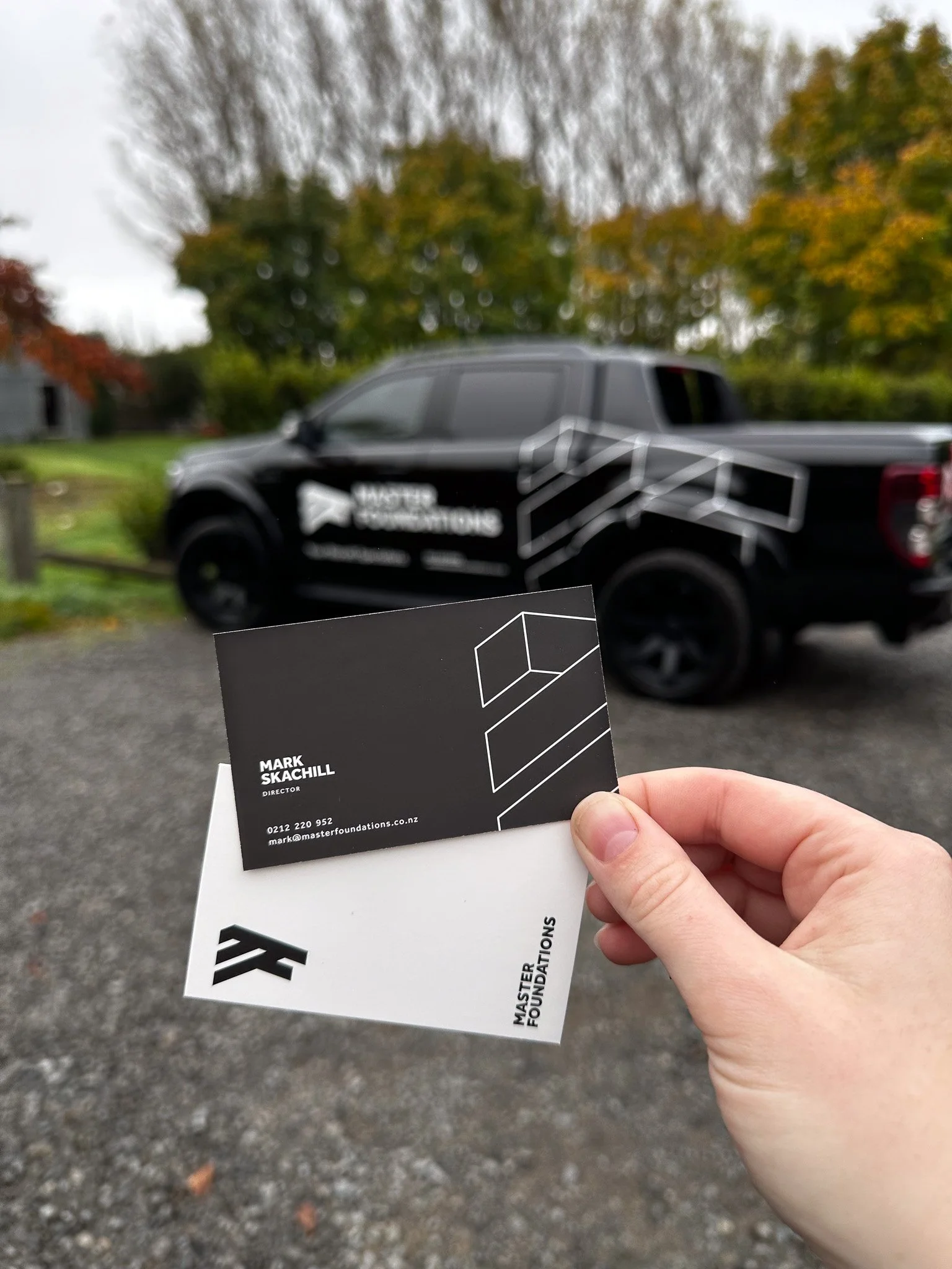 Person holding business cards in front of a black truck with branding for Master Foundations, parked outdoors with autumn trees in the background.