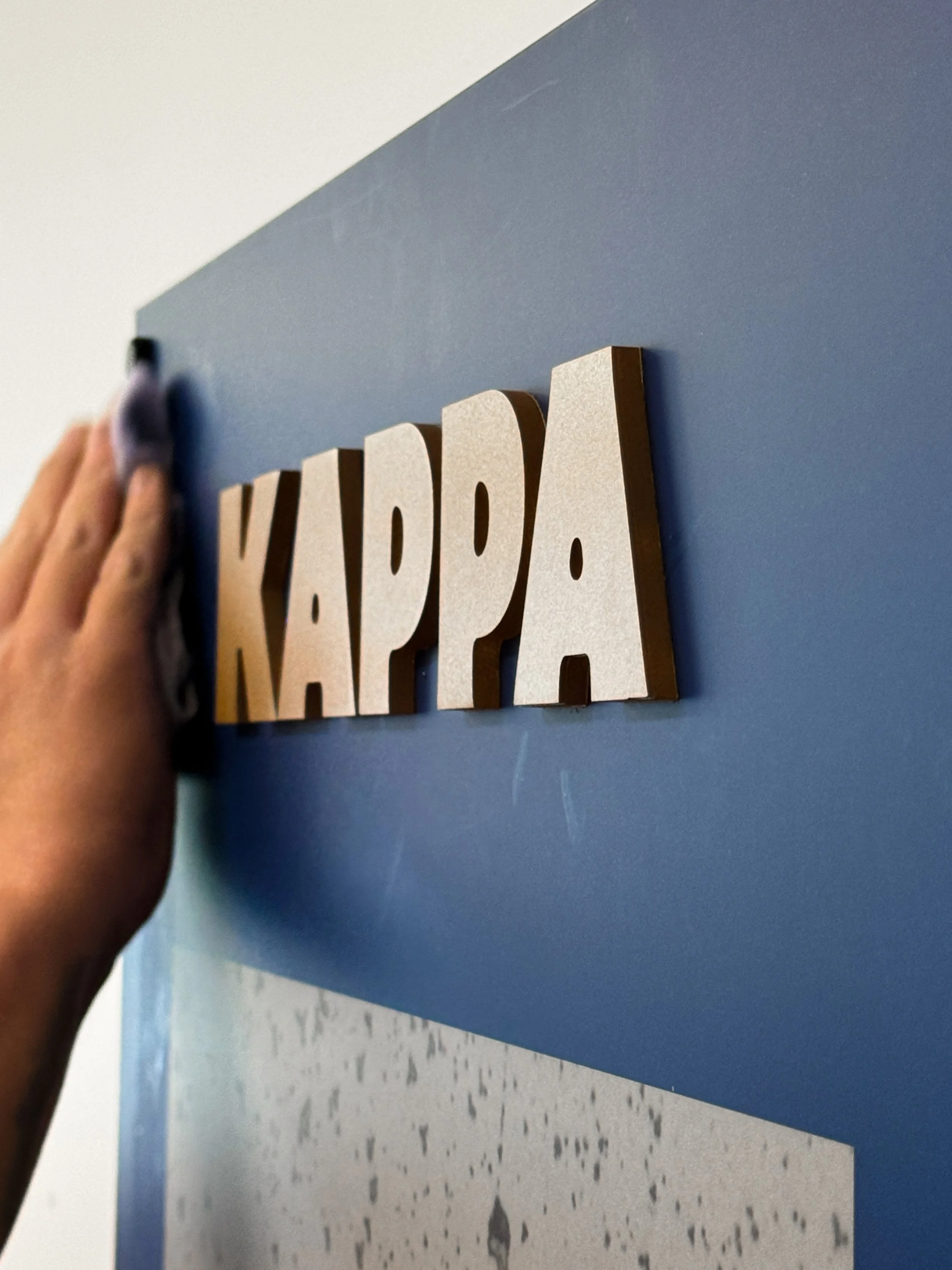Close-up of a person placing or adjusting a wooden sign that reads 'KAPPA' on a blue wall.
