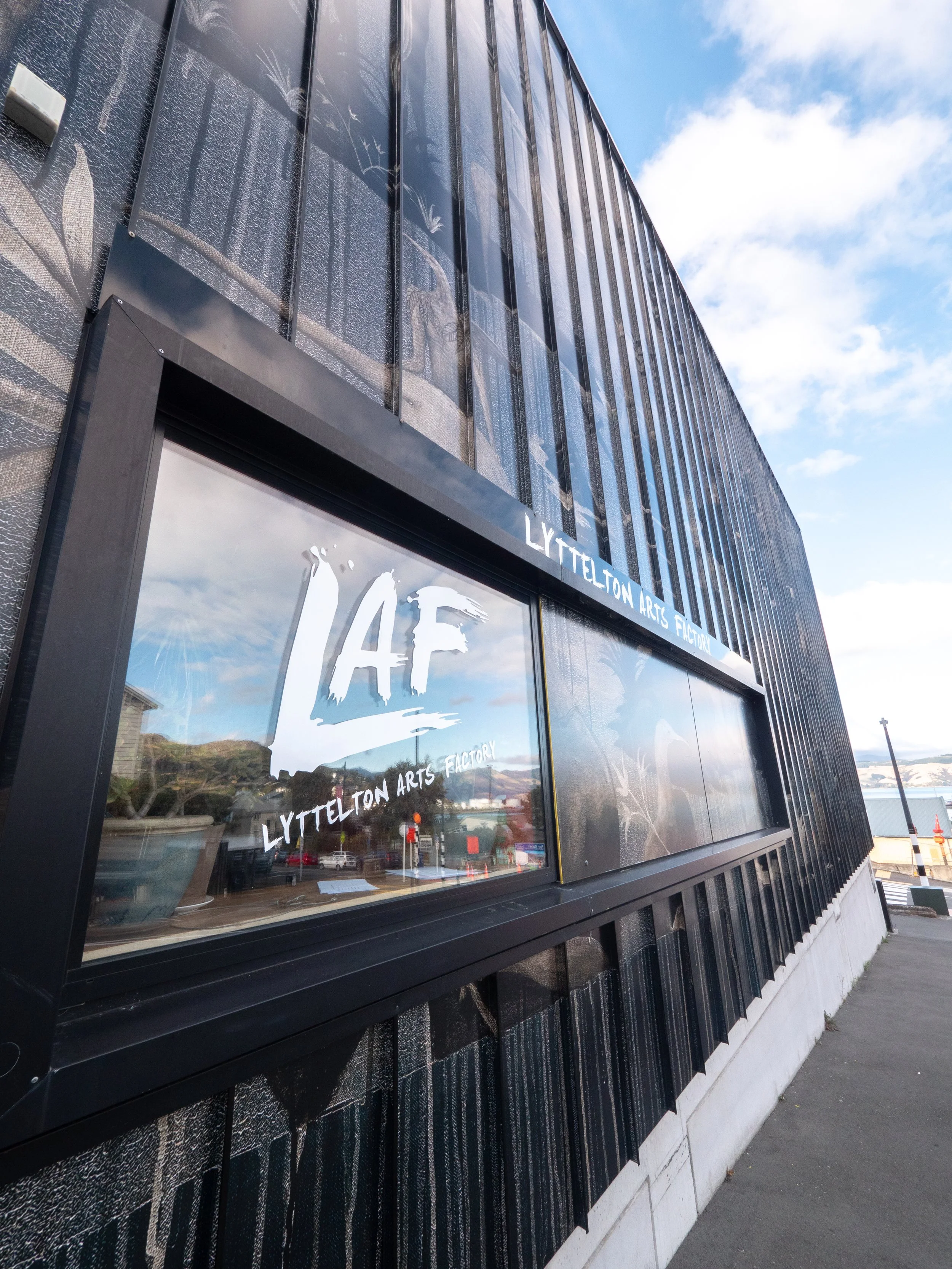 Exterior of a modern black building with large glass windows and white text reading "Lyttelton Arts Factory" on the window, under a partly cloudy sky.