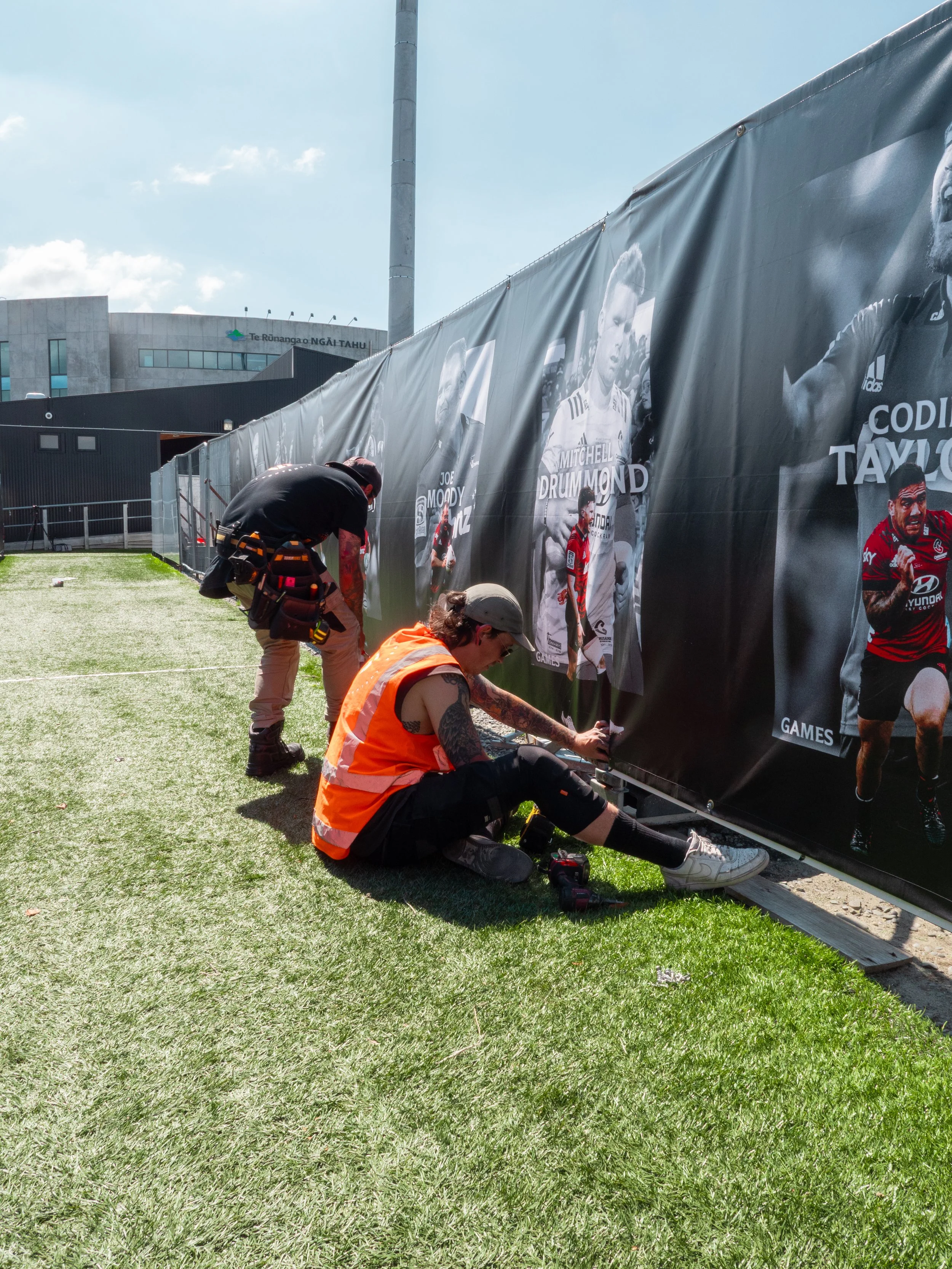 A woman with tattoos, wearing an orange safety vest and gray cap, kneels on the grass and attaches a poster to a large black banner with photos of athletes. A man in black with tools stands nearby, working on the banner. The scene is outdoors with a modern building in the background and a blue sky.