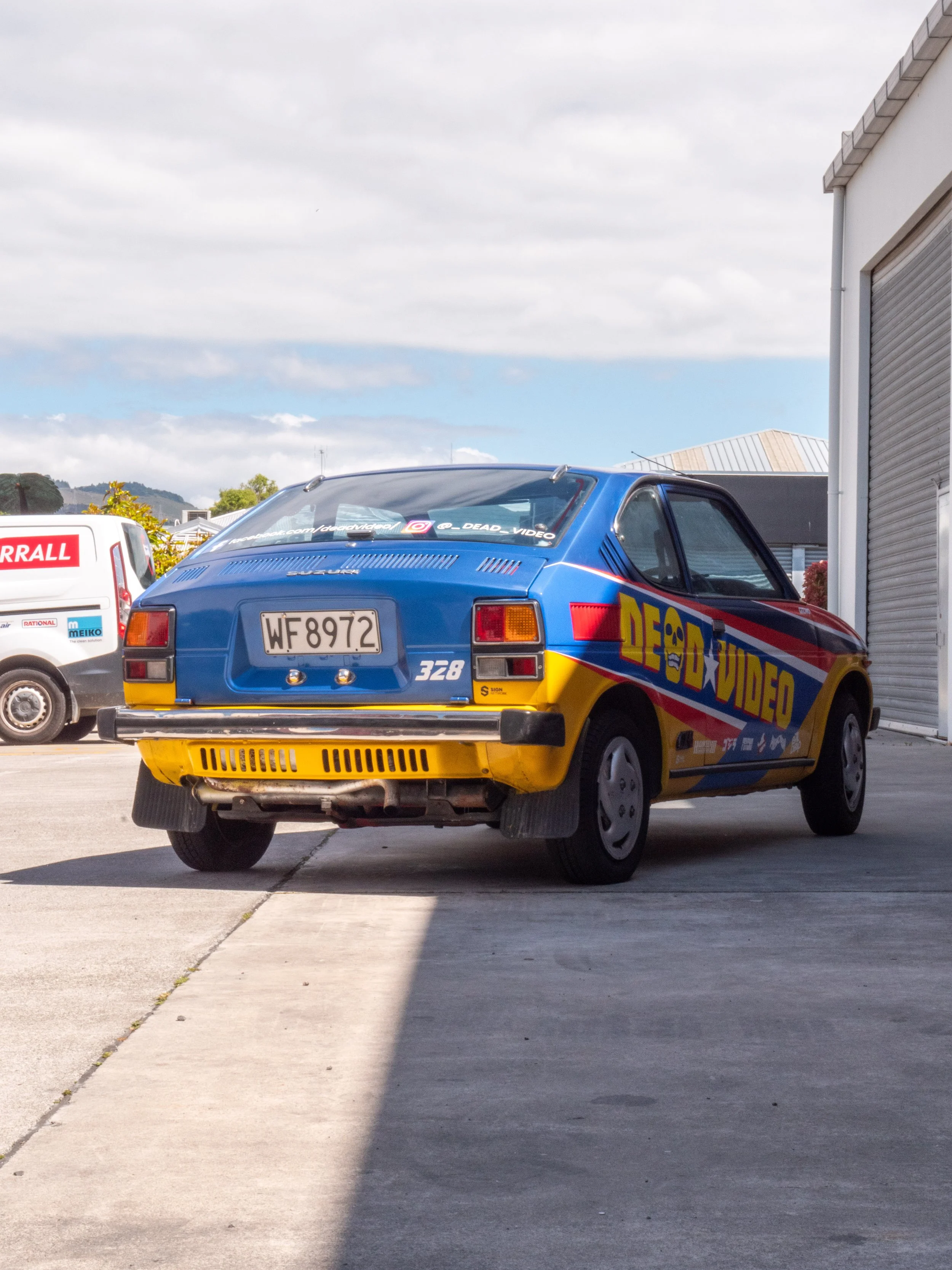 A vintage blue and yellow car with a 'DED VIDEO' logo parked on a concrete driveway beside a building, with a white van in the background against a partly cloudy sky.