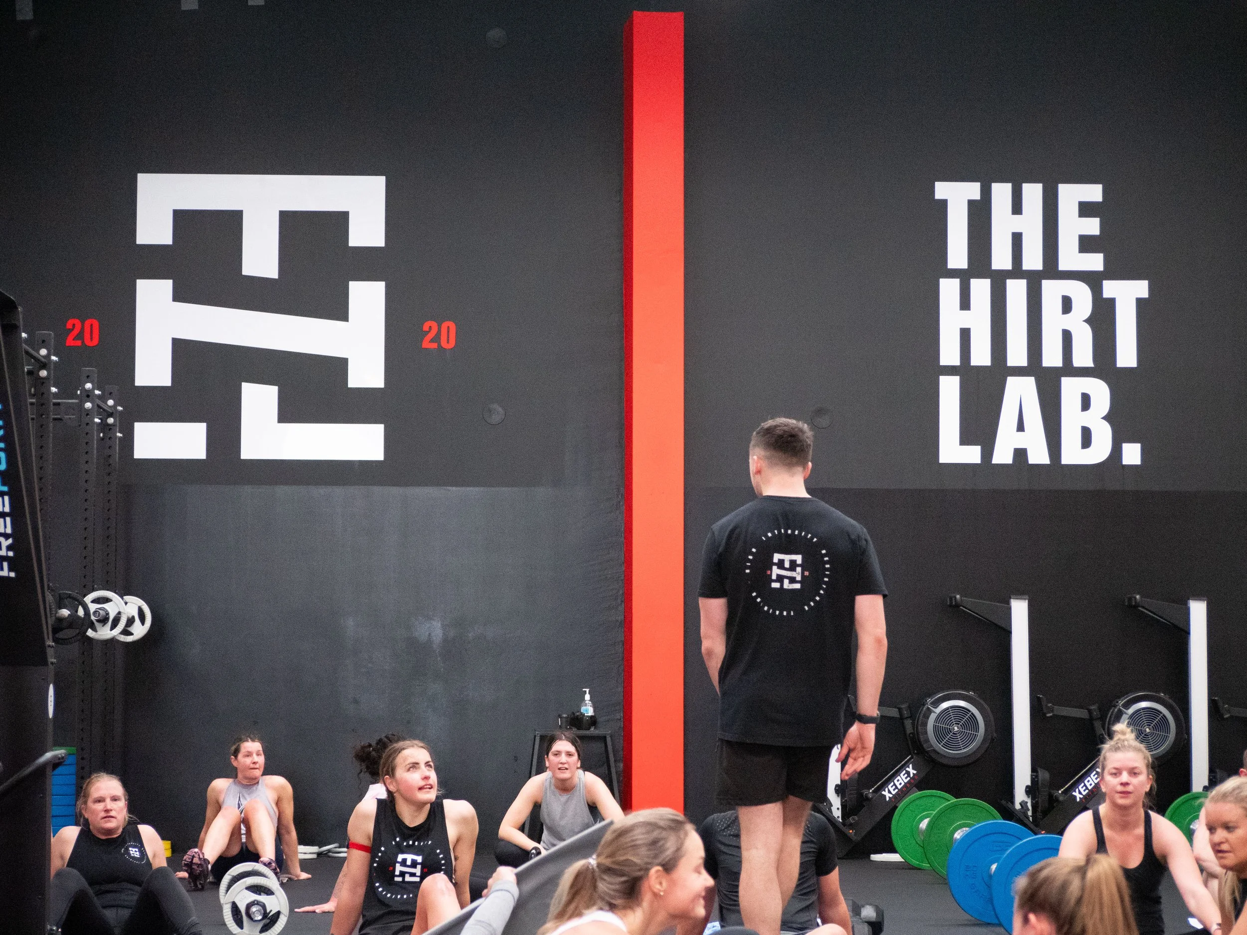 Group of women sitting on the floor at a fitness class in The Hirt Lab gym, with a trainer standing in front of them. The gym has black walls with large white text and a red stripe.
