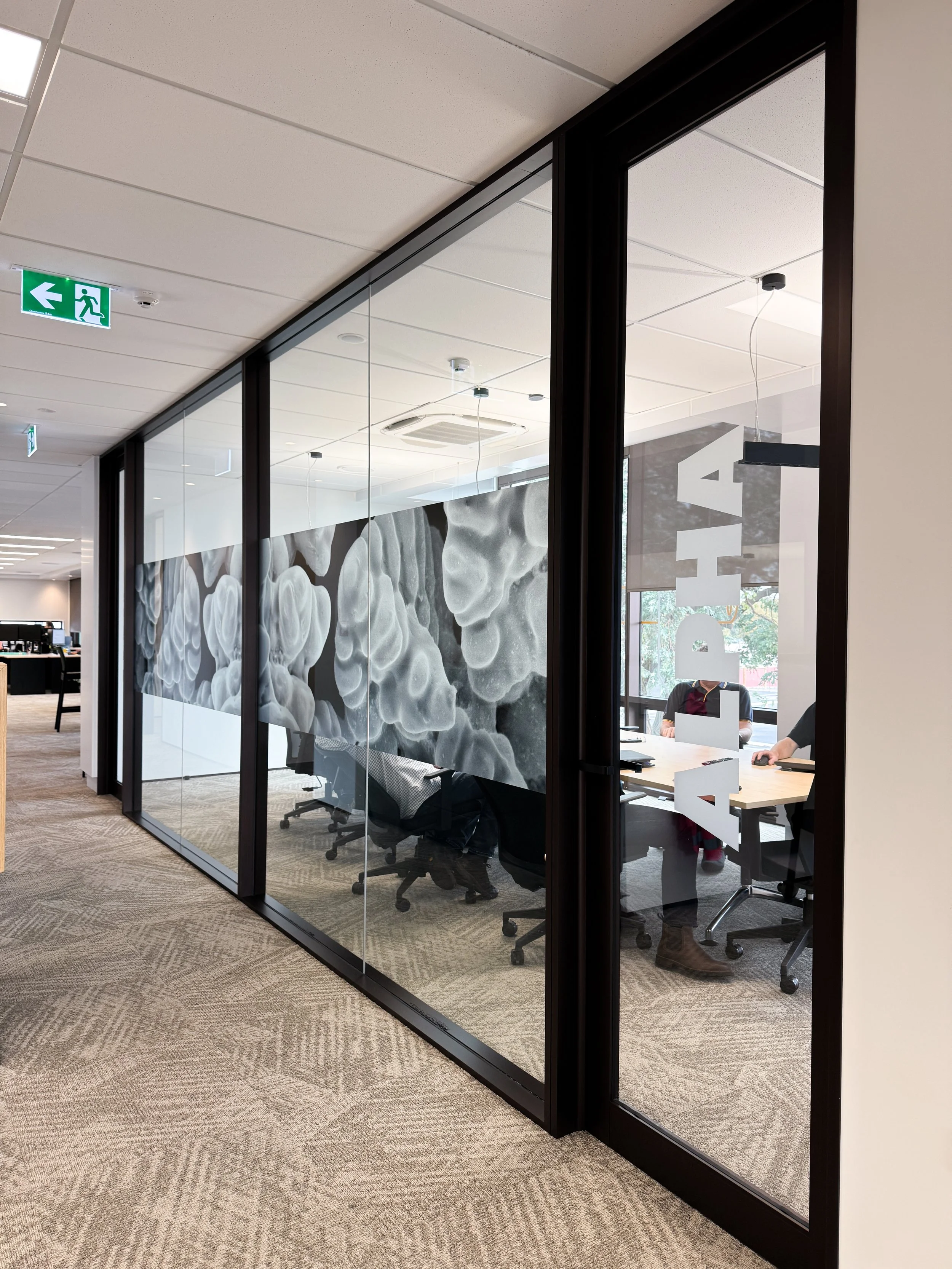 A glass-walled conference room with a large artistic black-and-white abstract mural on the back wall, a table with chairs around it, and two people sitting inside at the table. The room has ceiling air conditioning units and a window showing some outdoor trees.