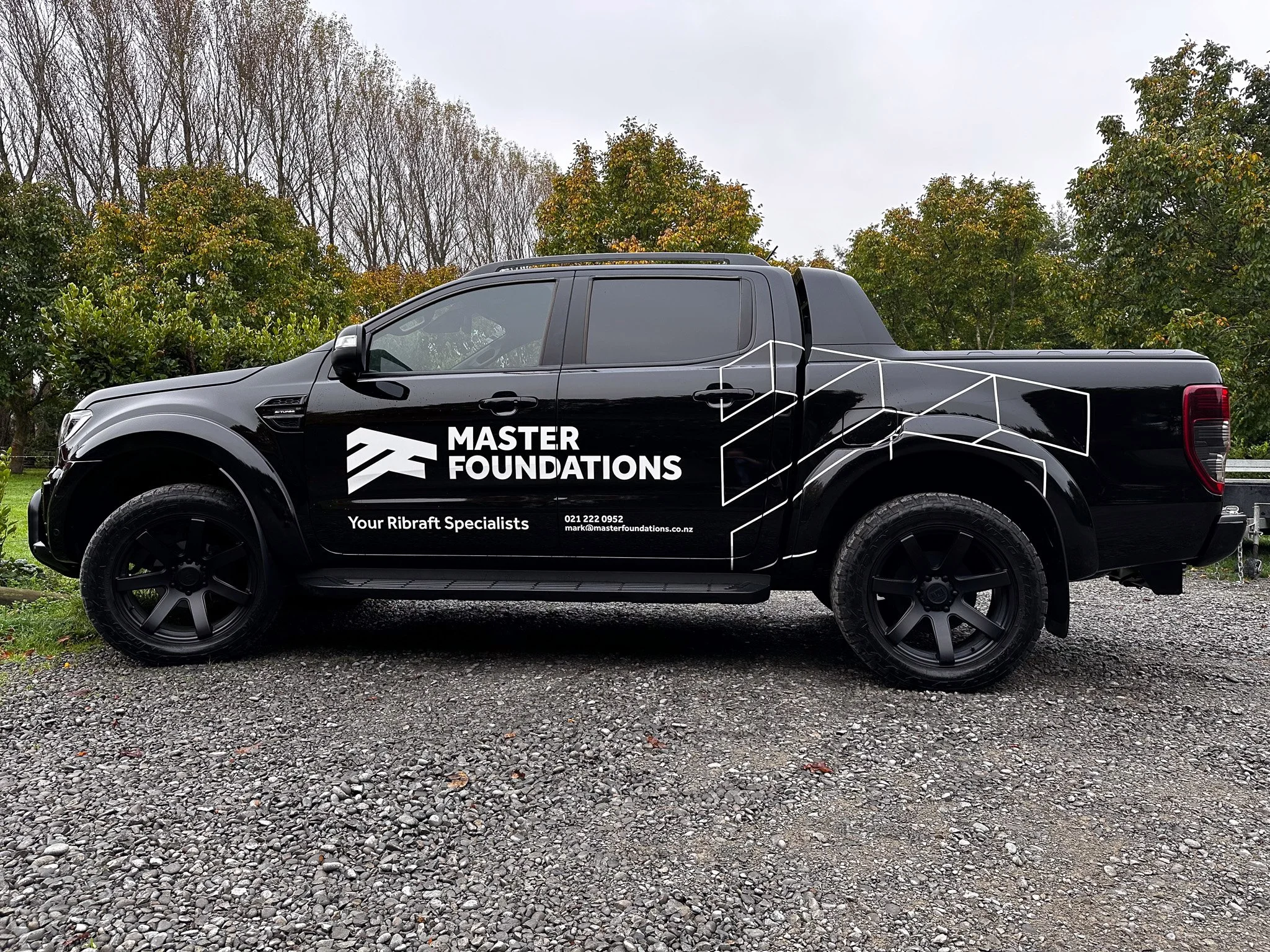 Black pickup truck with company branding for Master Foundations, displaying contact information and slogan 'Your Ribraft Specialists', parked on a gravel surface with trees in the background.