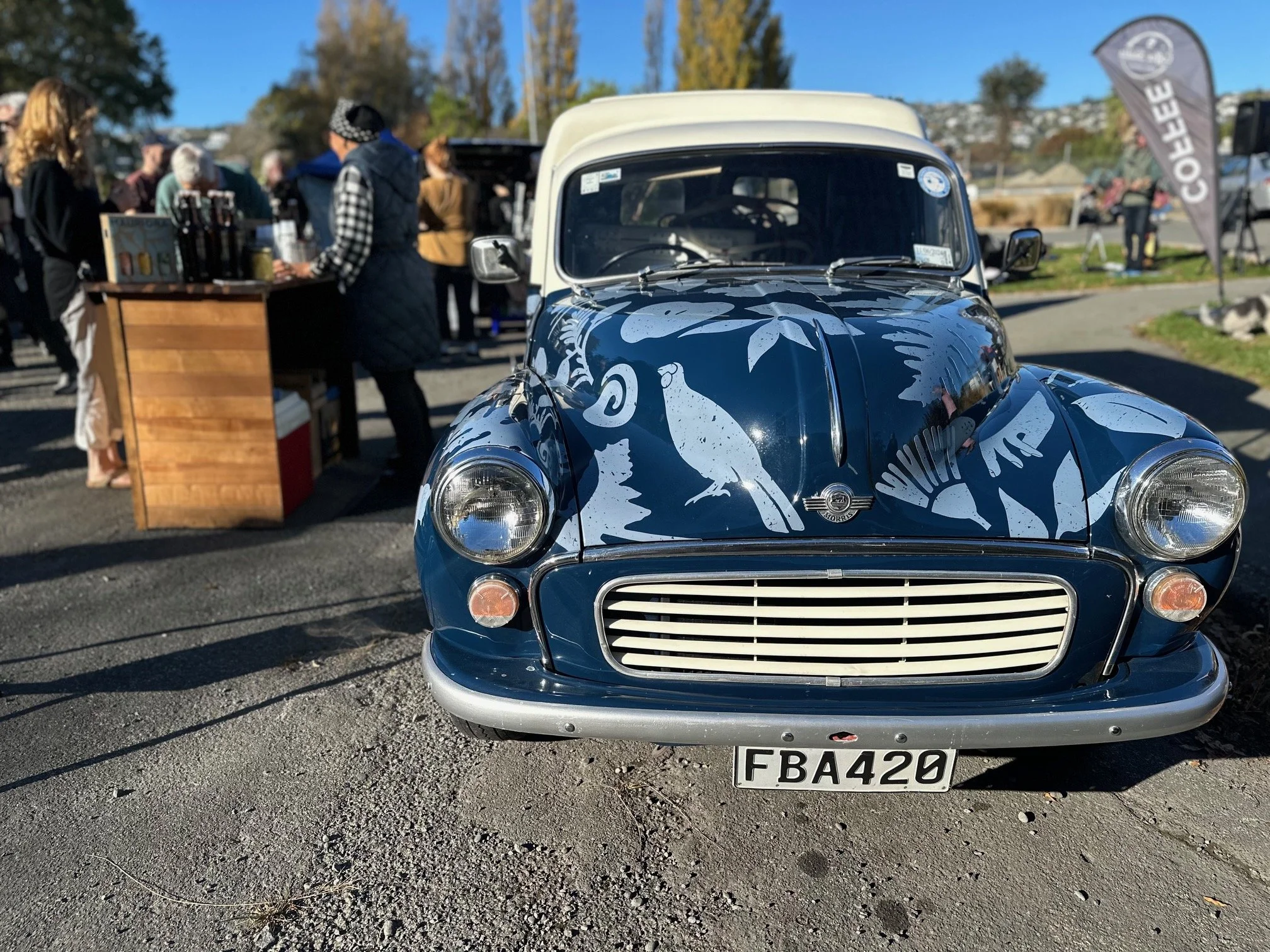 A vintage blue car with white leaf and bird illustrations, parked at an outdoor event with people and vendor stalls in the background.