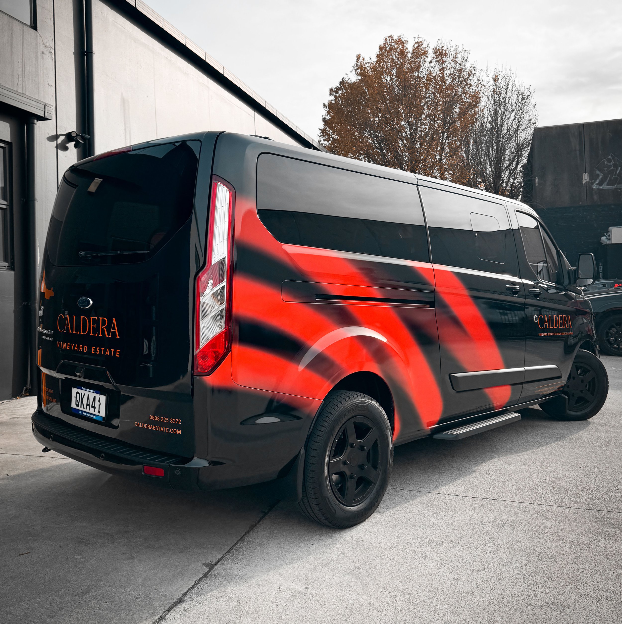 Black van with red and black curved design and "Caldera Vineyard Estate" logo parked outside a building with trees in the background.