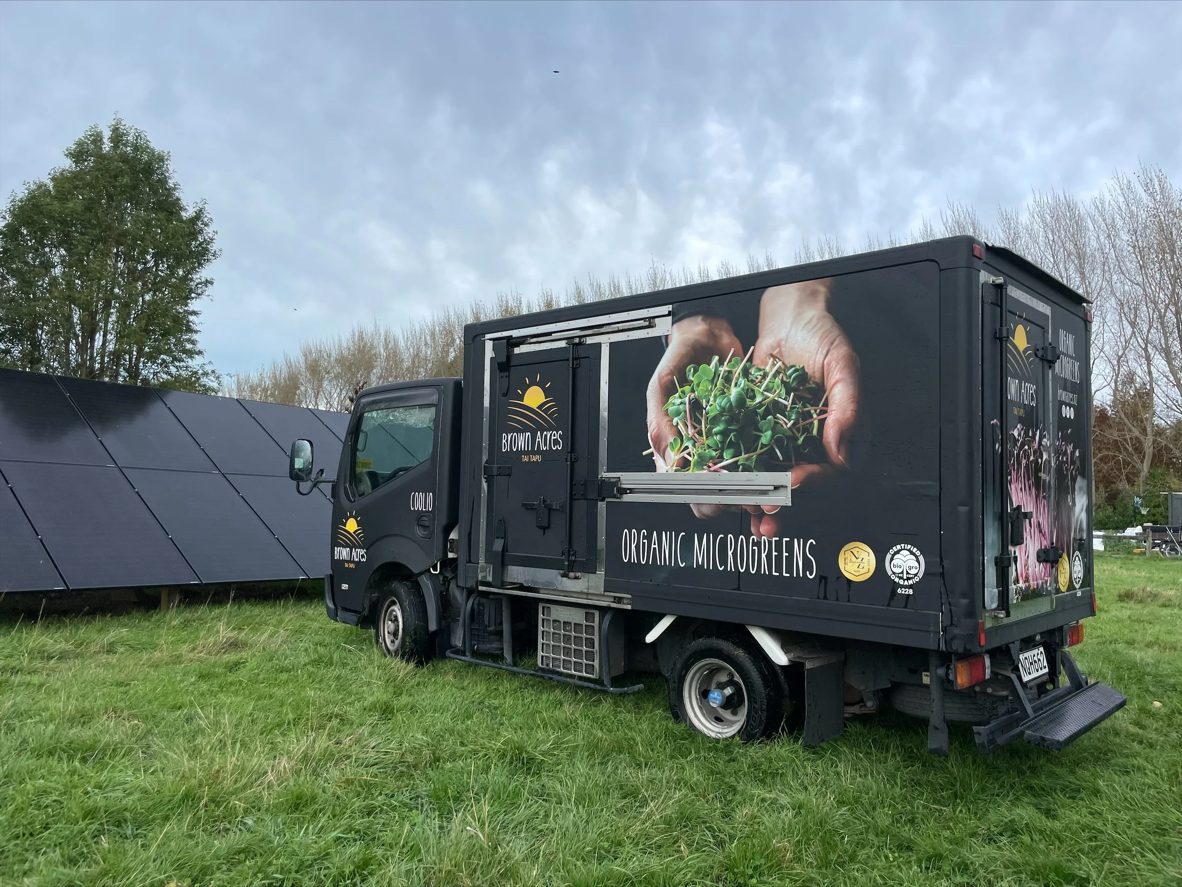A black food truck with graphics advertising organic microgreens, parked on a grassy field with solar panels nearby and trees in the background.