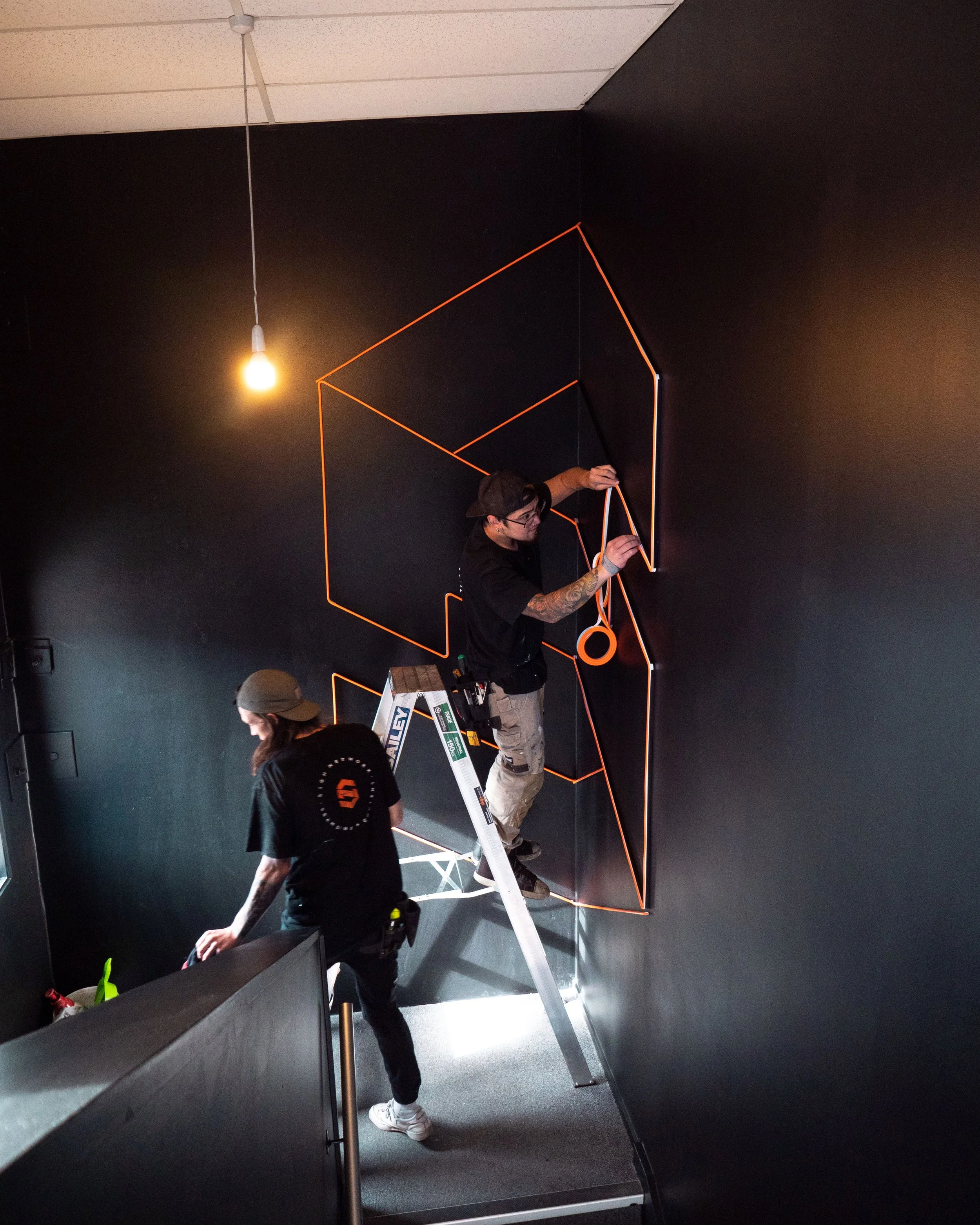 Two workers installing orange neon light strips on a black wall, one on a ladder and one standing nearby.