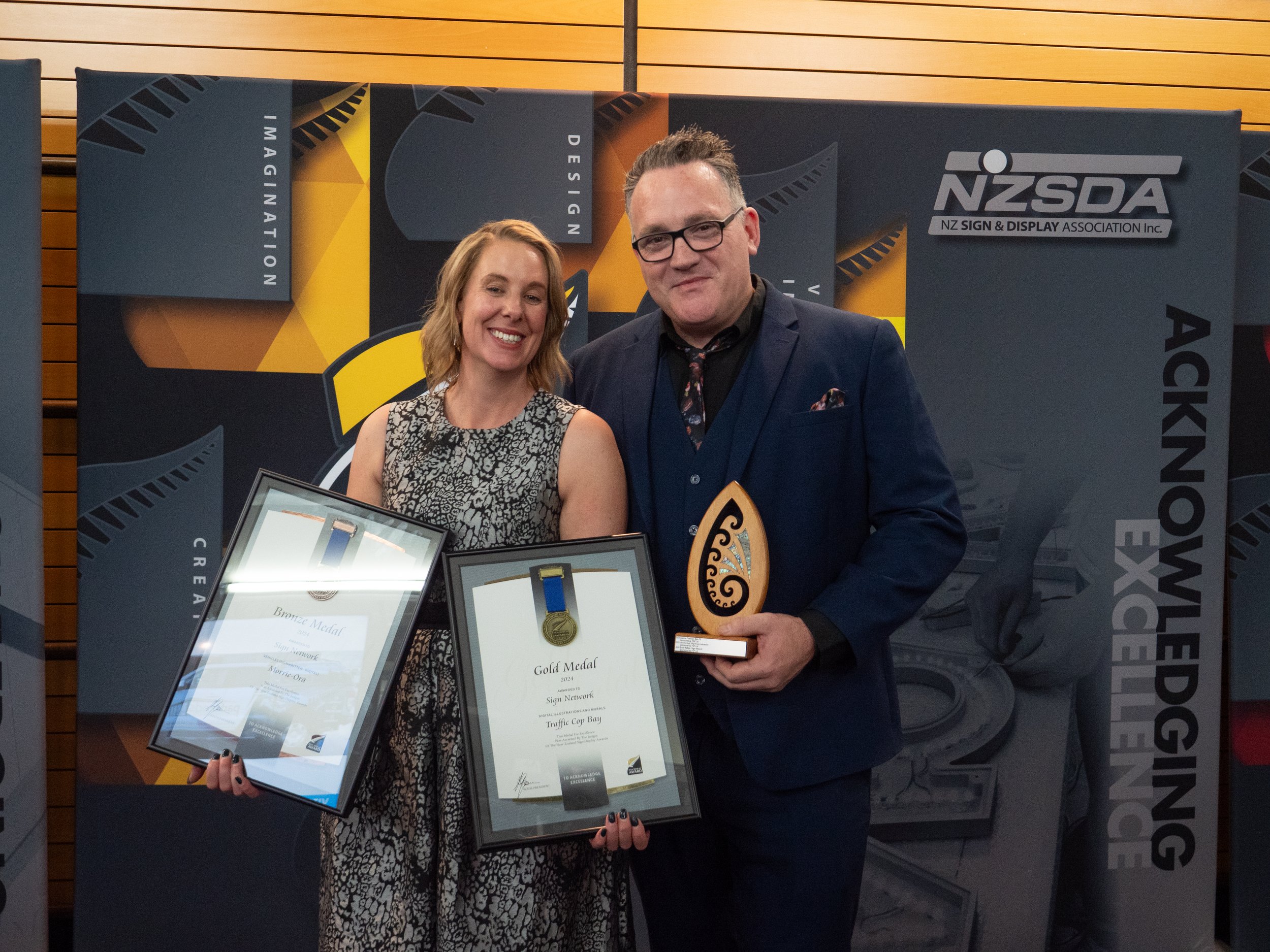Two people, a woman and a man, standing together at an awards ceremony. The woman is holding two framed certificates, and the man is holding a trophy. They are smiling and dressed in formal attire. The background features a banner with the NZSDA logo and words like "Design," "Creative," and "Imagine".