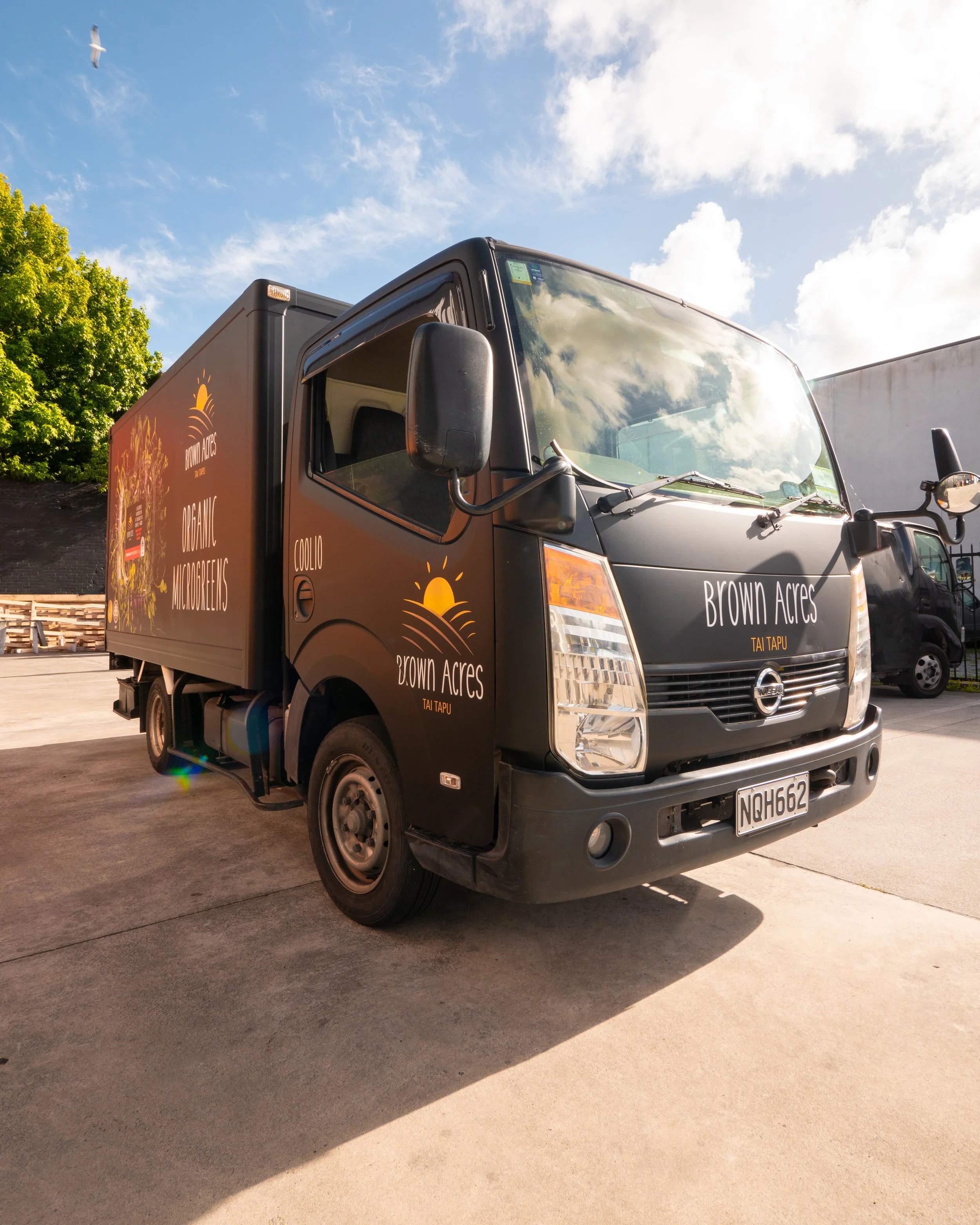 Black delivery truck with 'Brown Acres' logo and text, parked on concrete, with blue sky and clouds overhead and a bird flying in the background.