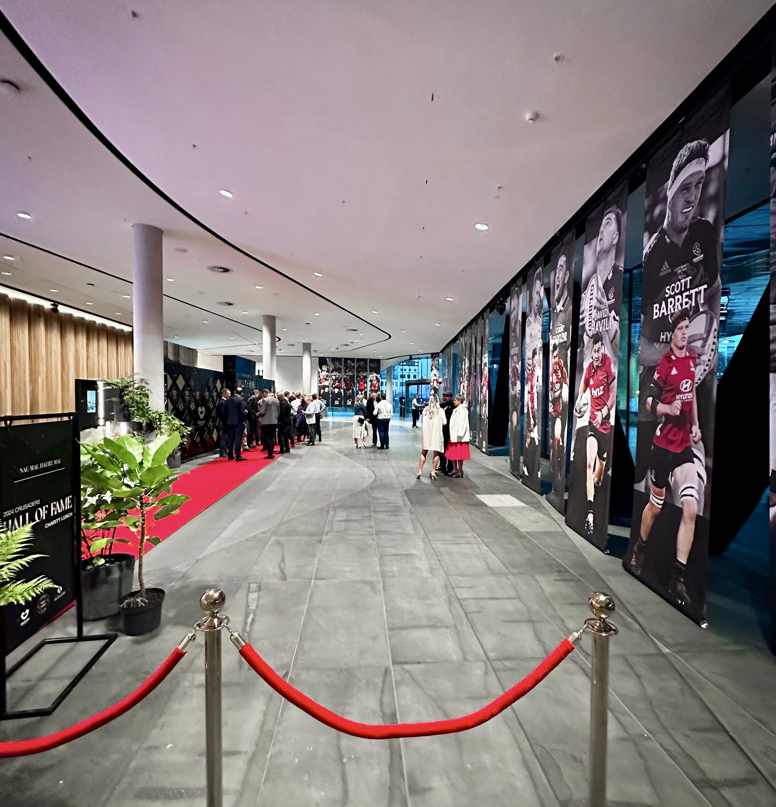 Indoor event with a group of people socializing, a red carpet on the left, and tall banners displaying athletic players on the right side of the hallway.