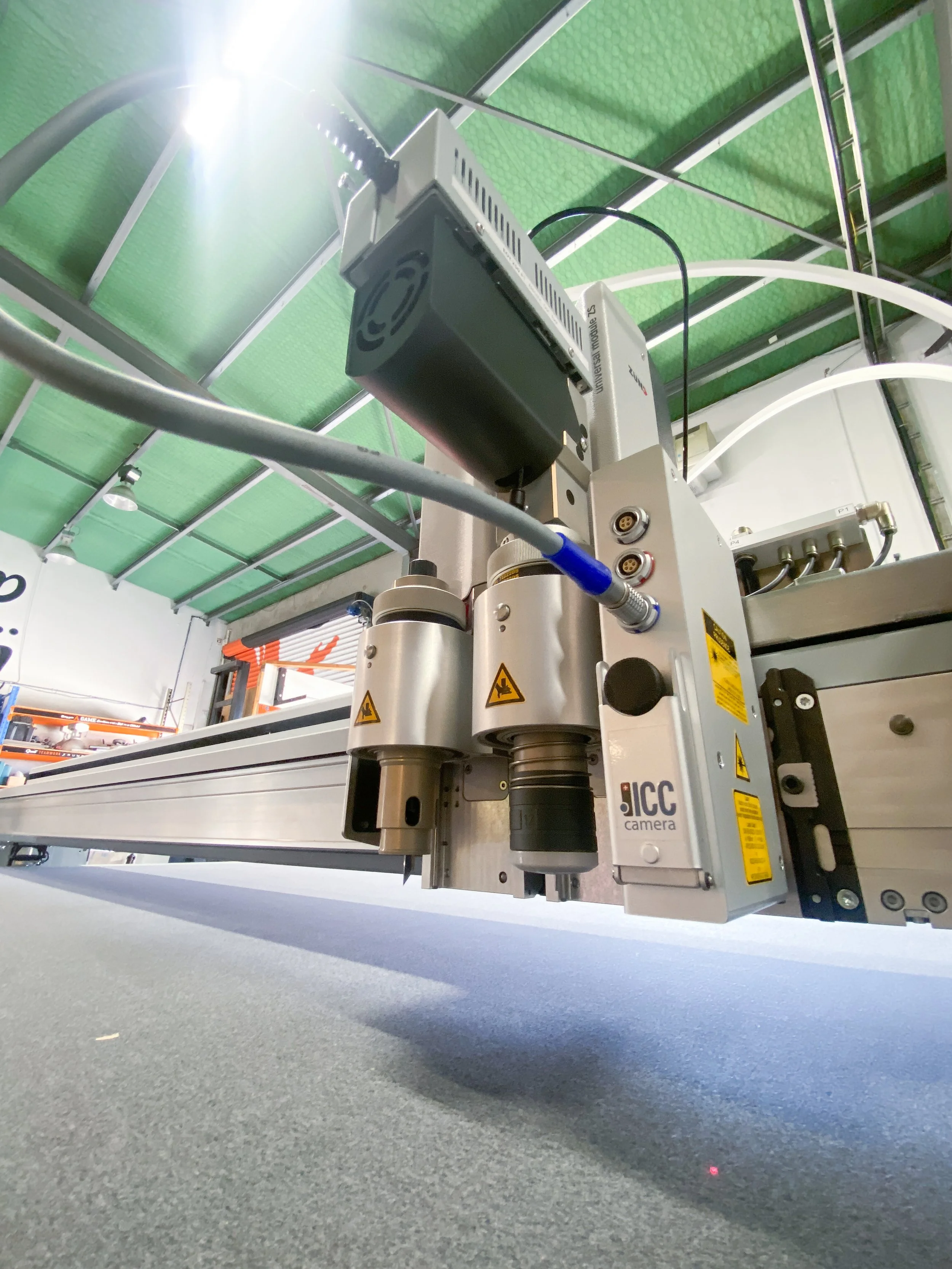 Close-up view of a CNC machine or industrial robotic equipment in a factory setting with green ceiling panels and industrial lighting.