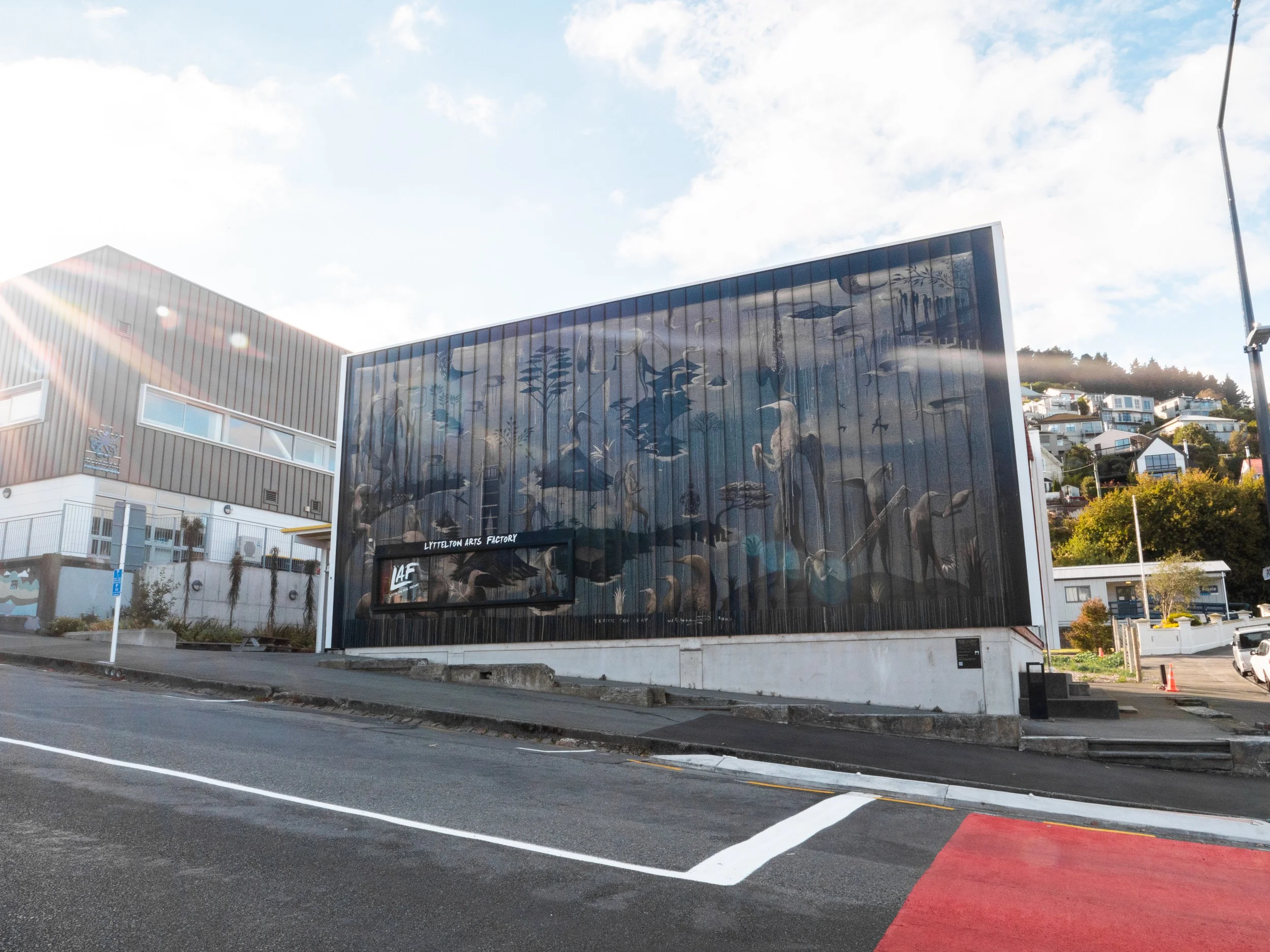 A modern building with a large mural featuring aquatic animals and birds, located in an urban setting with houses on a hillside in the background. The building has black exterior walls and a sign reading "Lyttelton Arts Factory."